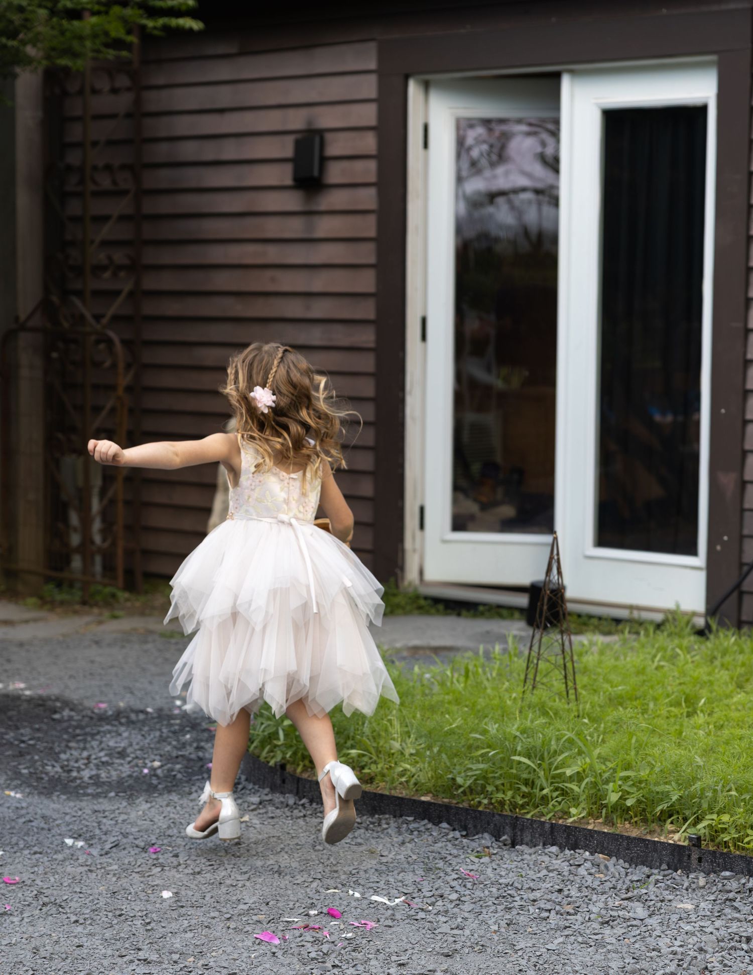Young flower girl in white tulle dress tosses confetti outside a dark wood building.