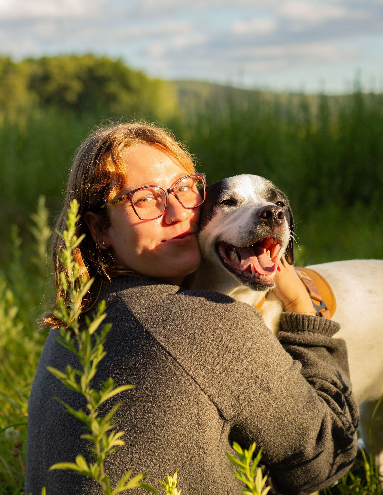 Smiling woman with glasses hugs happy white dog in sunny green meadow.