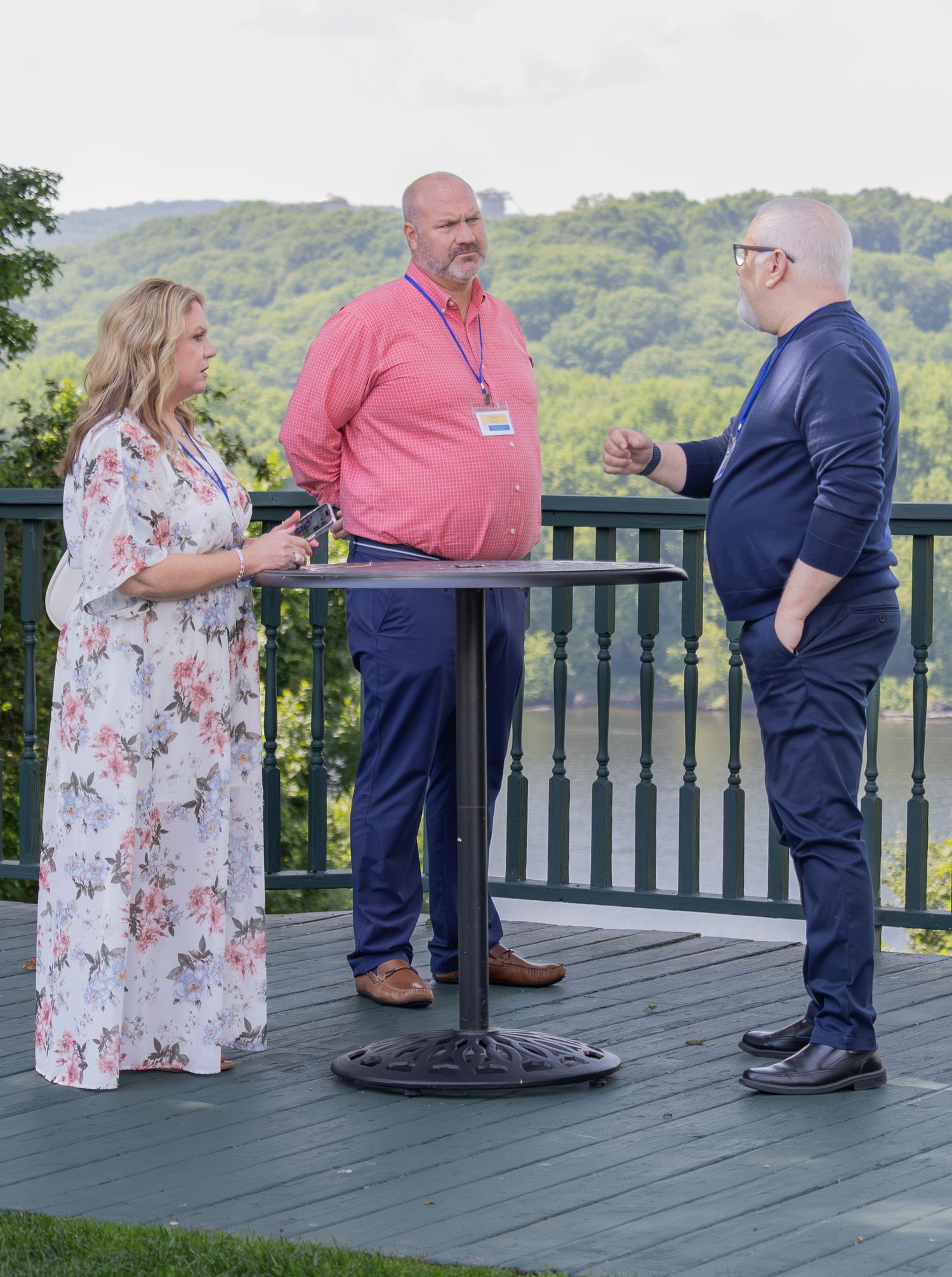 Three professionals networking on outdoor deck with scenic green hillside background at corporate event.