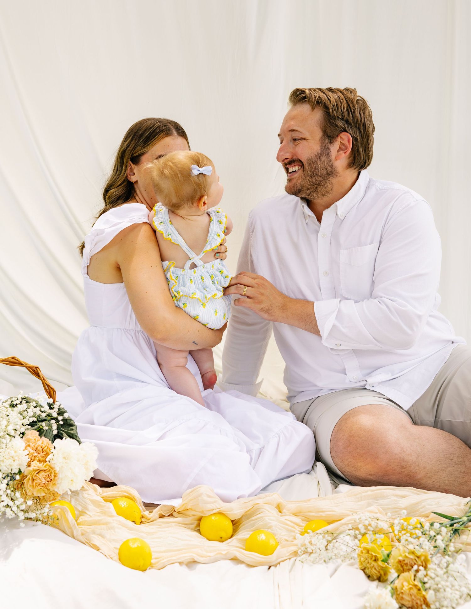 Happy parents in white outfits laughing with their baby among lemons and flowers.