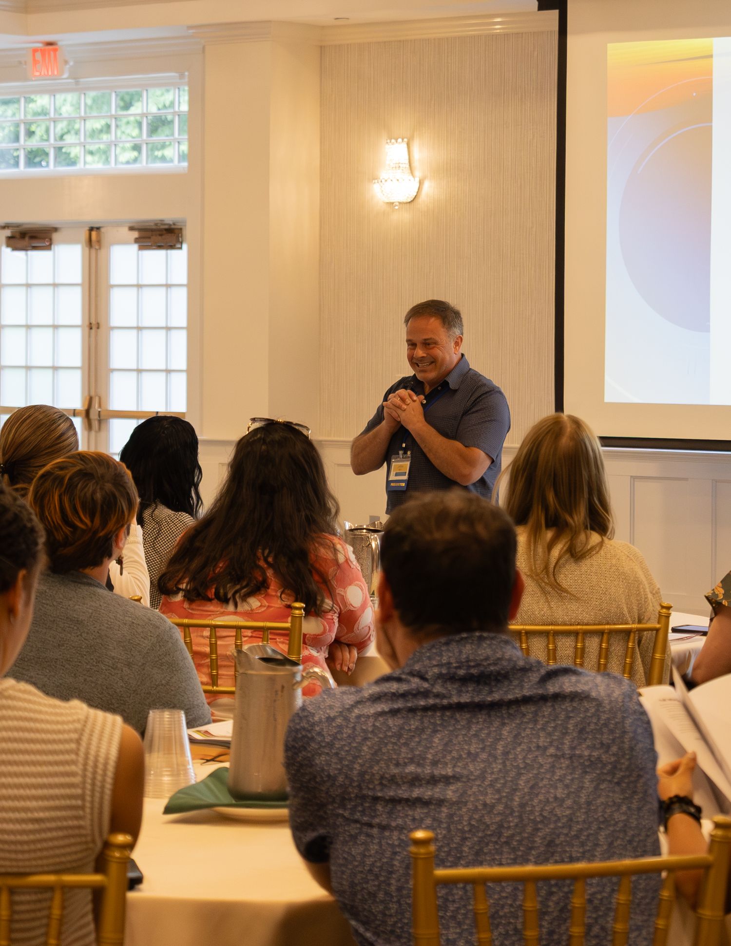 Male speaker presenting to a group of attendees seated at tables in a bright conference room.
