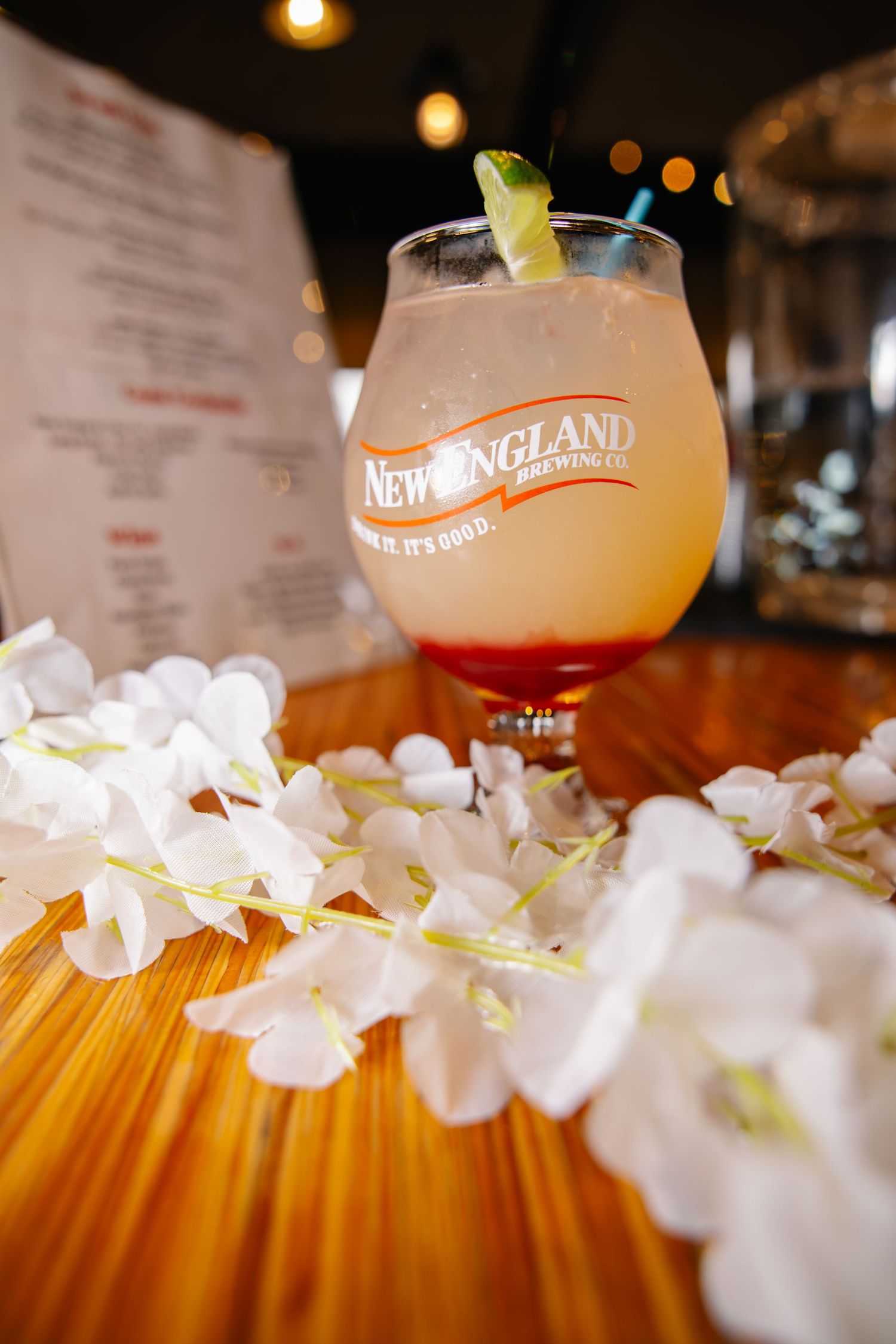 New England Brewing Co cocktail garnished with cucumber surrounded by white flowers on wooden bar.
