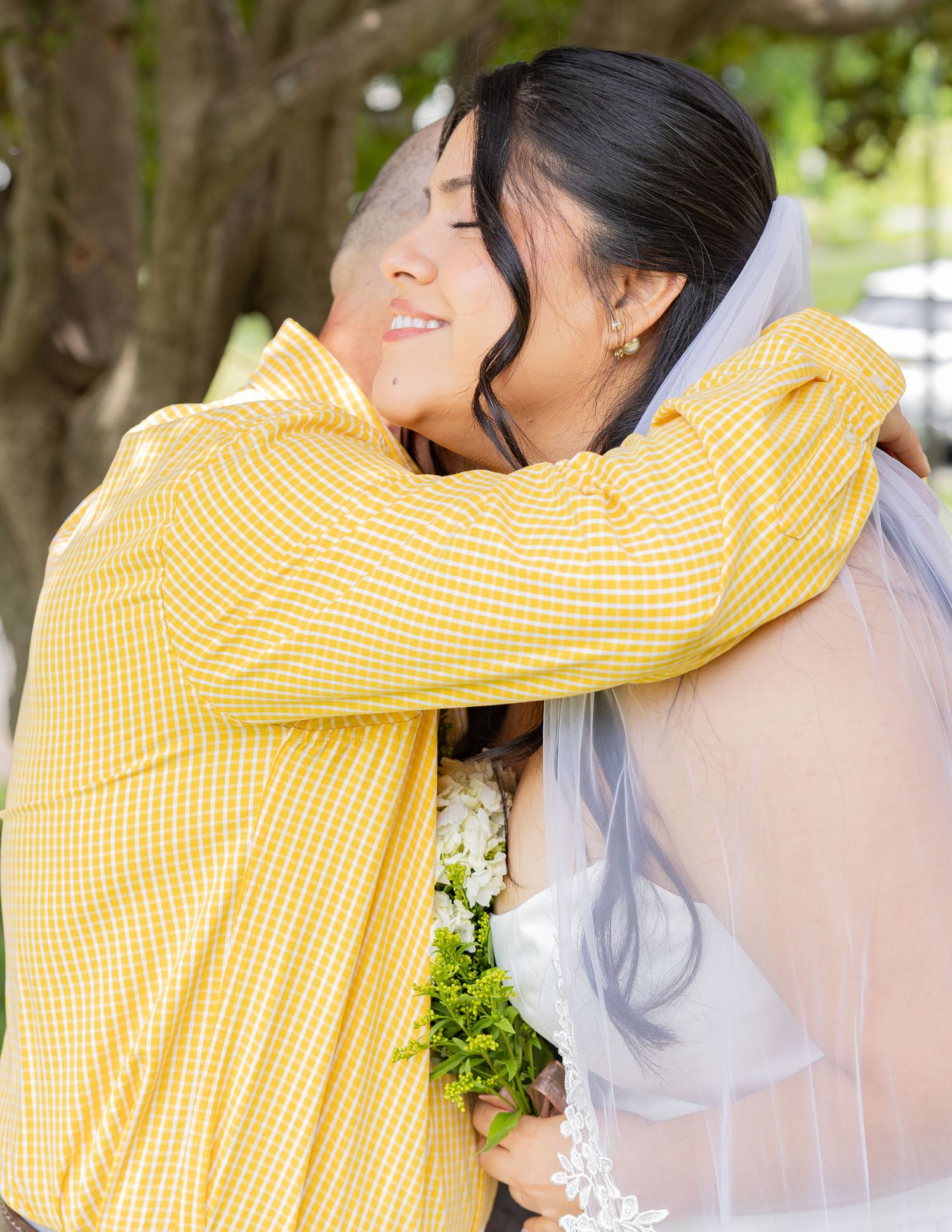 Bride in white gown and veil embracing father in yellow shirt under shaded outdoor setting.