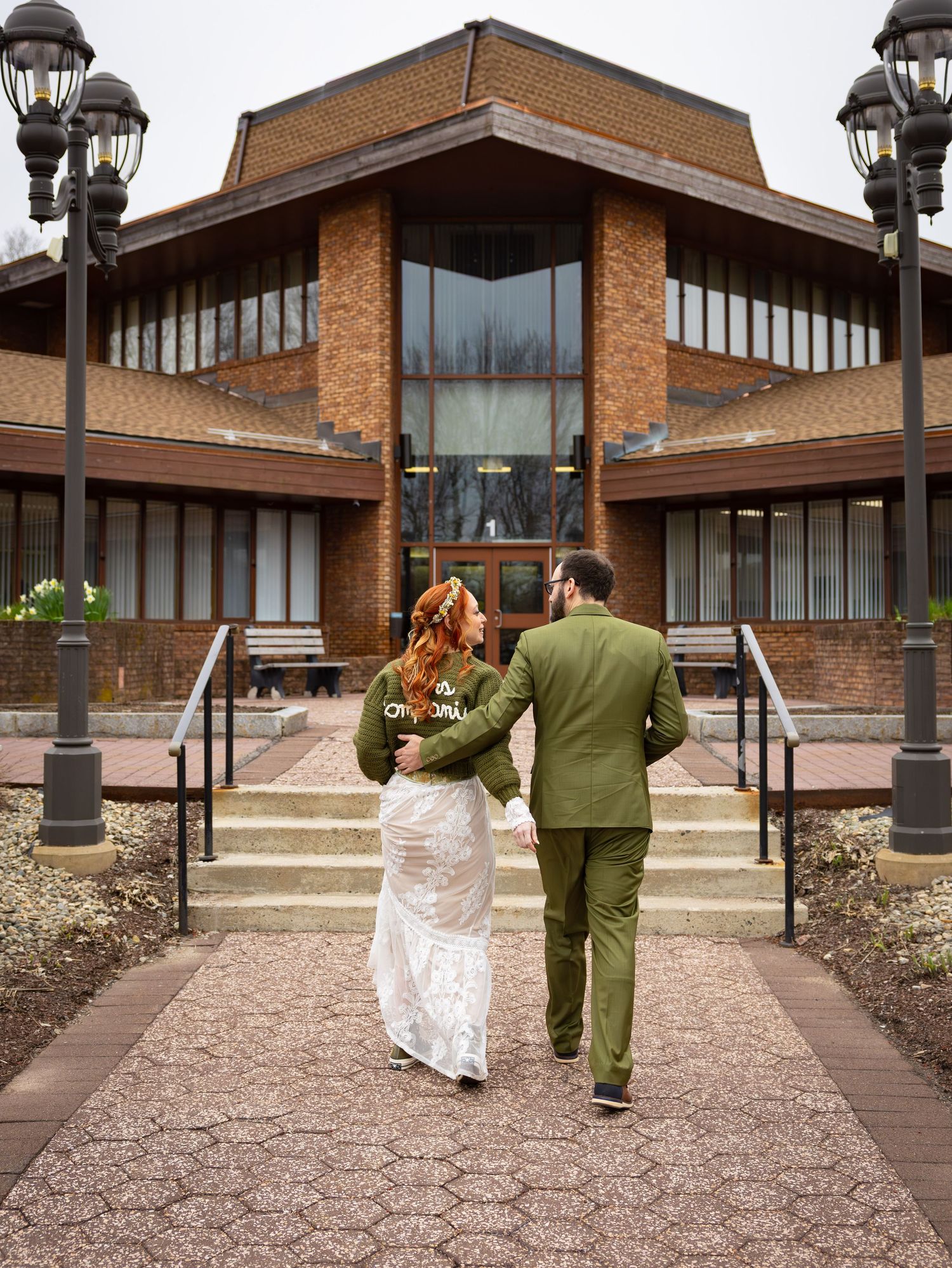 Newlywed couple walks toward brick building, bride in white dress groom in olive green suit.