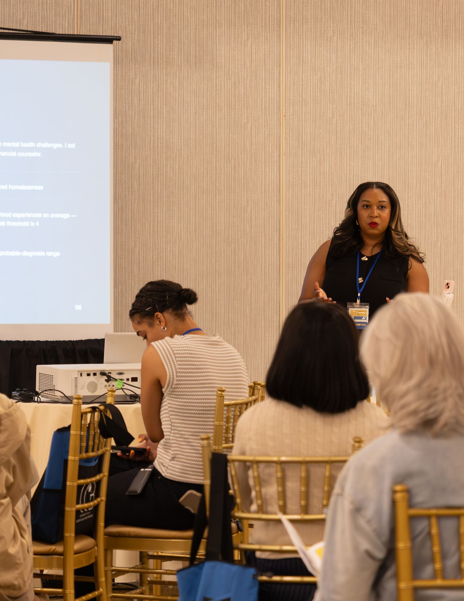 Woman speaker presents to seated audience at professional conference in hotel ballroom setting.