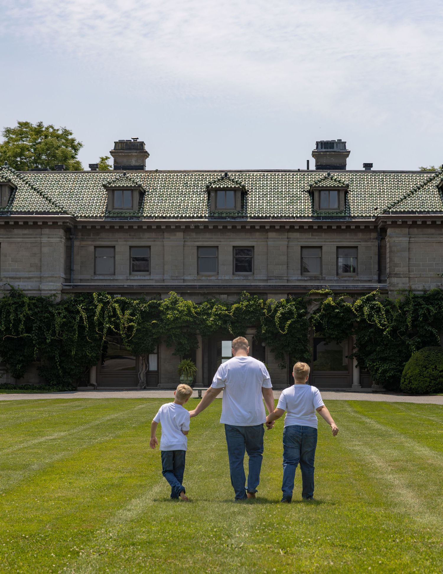 Father holding hands with two young sons walking toward grand historic mansion on sunny day.