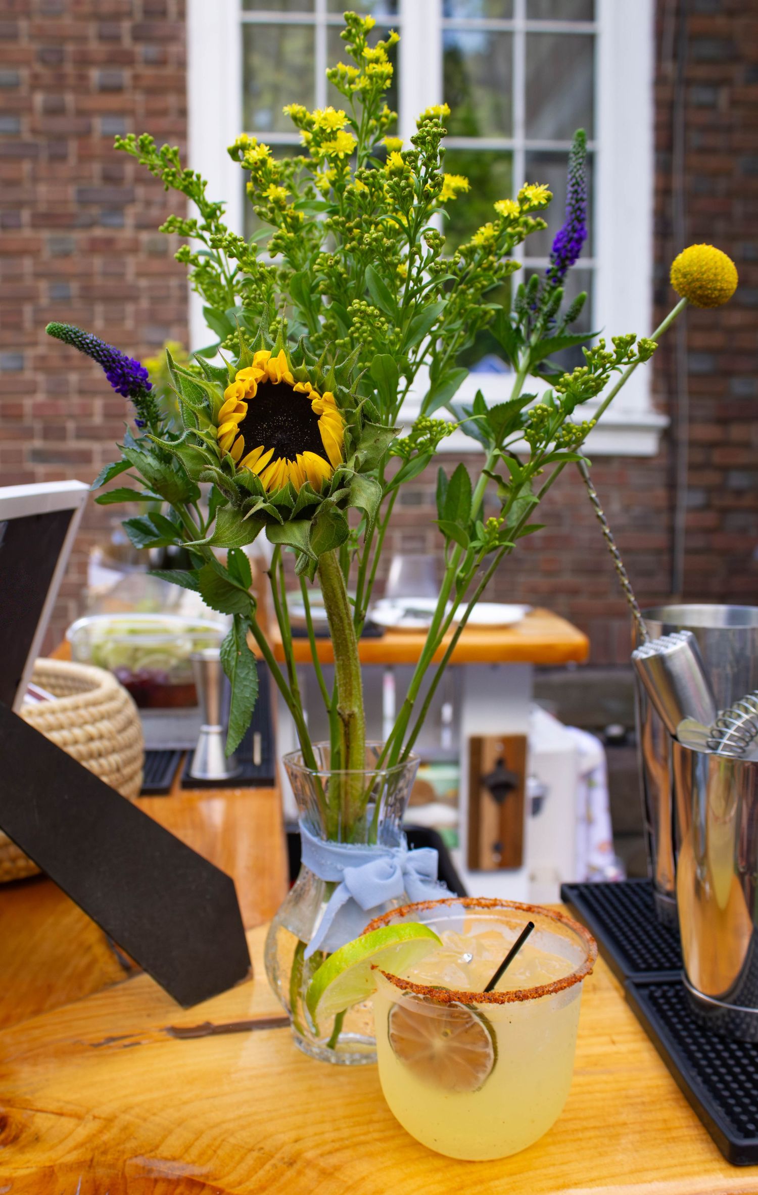 Wildflower bouquet with sunflower in glass vase beside citrus cocktail on outdoor bar table.