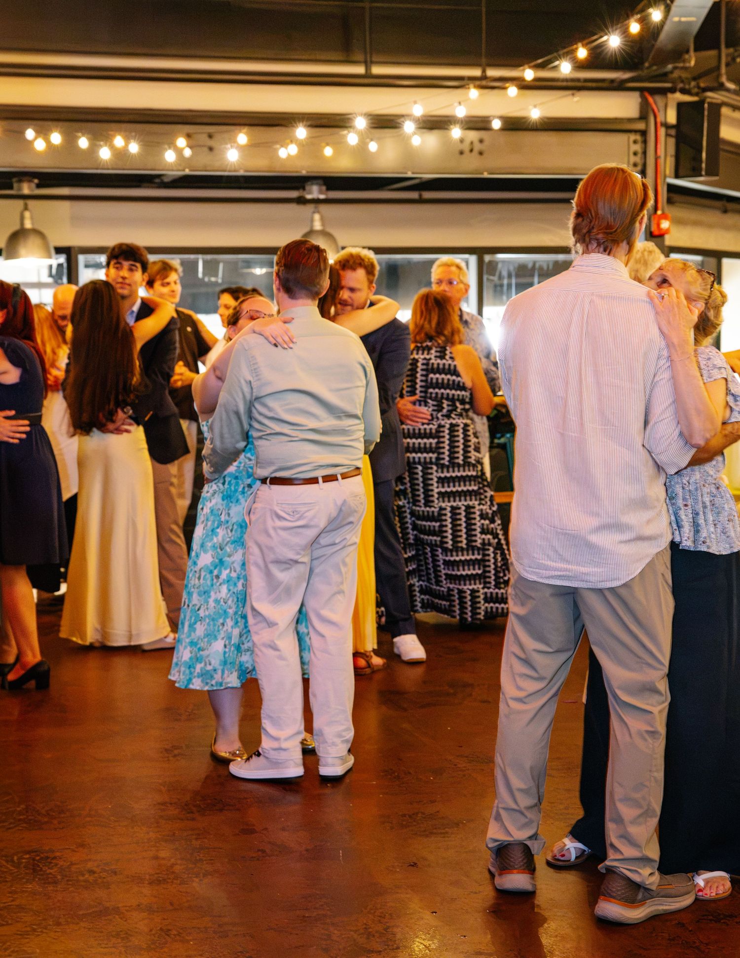 Guests slow dancing together at an indoor wedding reception with warm string lights overhead.