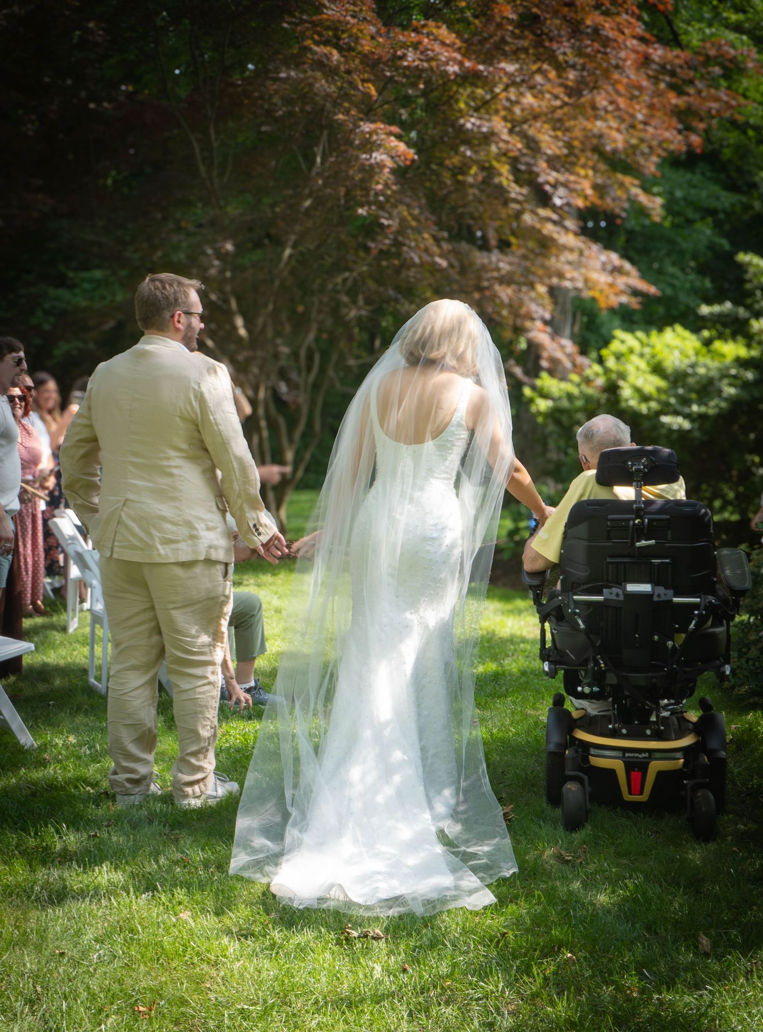 Bride in white gown and veil walks down outdoor aisle with groom, guest in motorized wheelchair nearby.