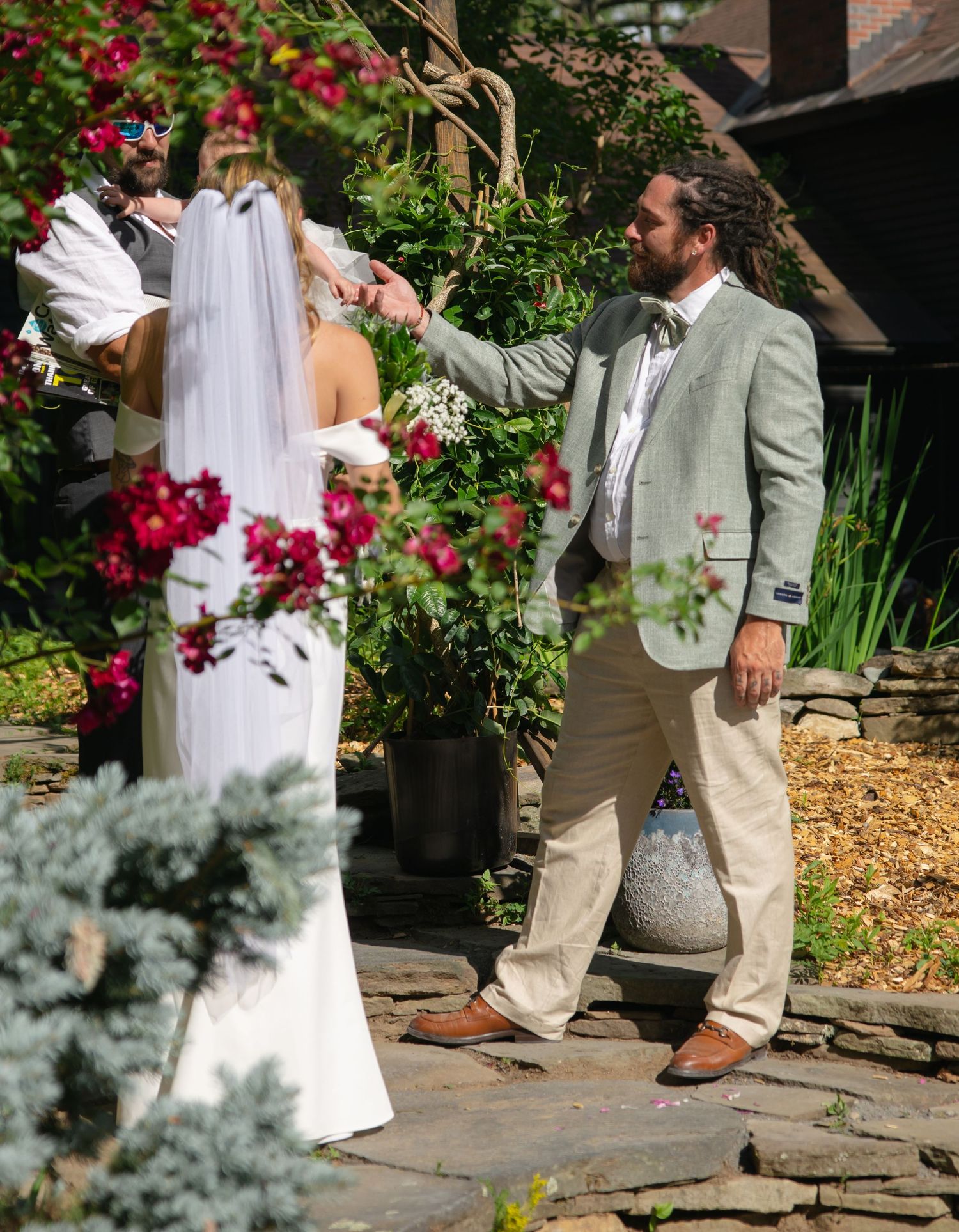 Groom in sage green suit holding bride's hand during outdoor garden wedding ceremony.