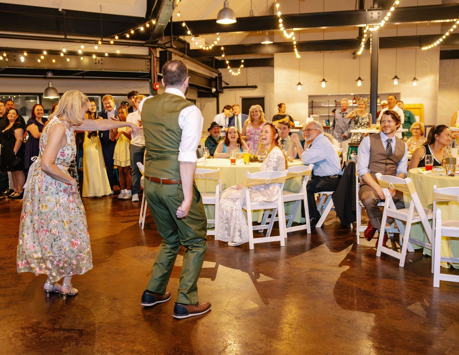 Wedding reception dance floor with guests dancing under string lights in bright industrial venue.