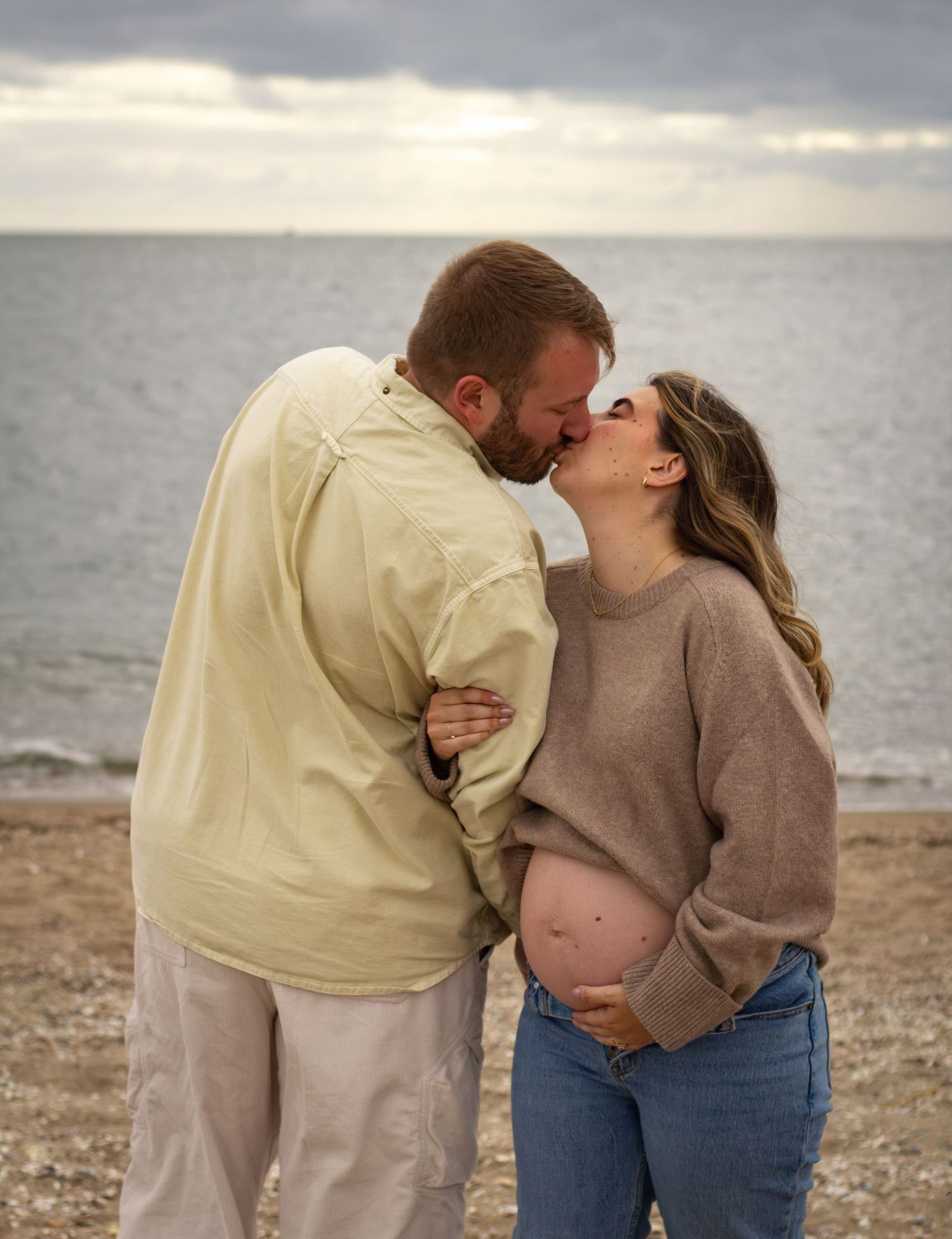 Pregnant couple sharing a kiss on a pebble beach with ocean backdrop on a cloudy day.
