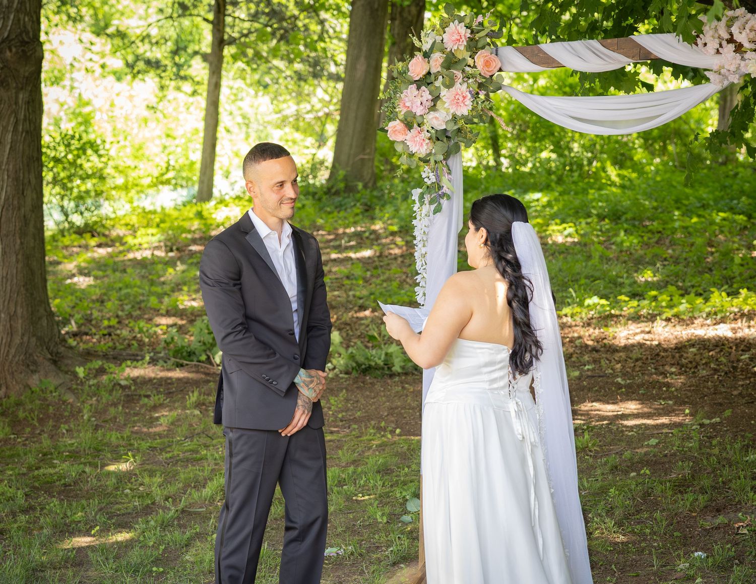Groom smiling at bride reading vows under a floral arch at an outdoor woodland wedding ceremony.