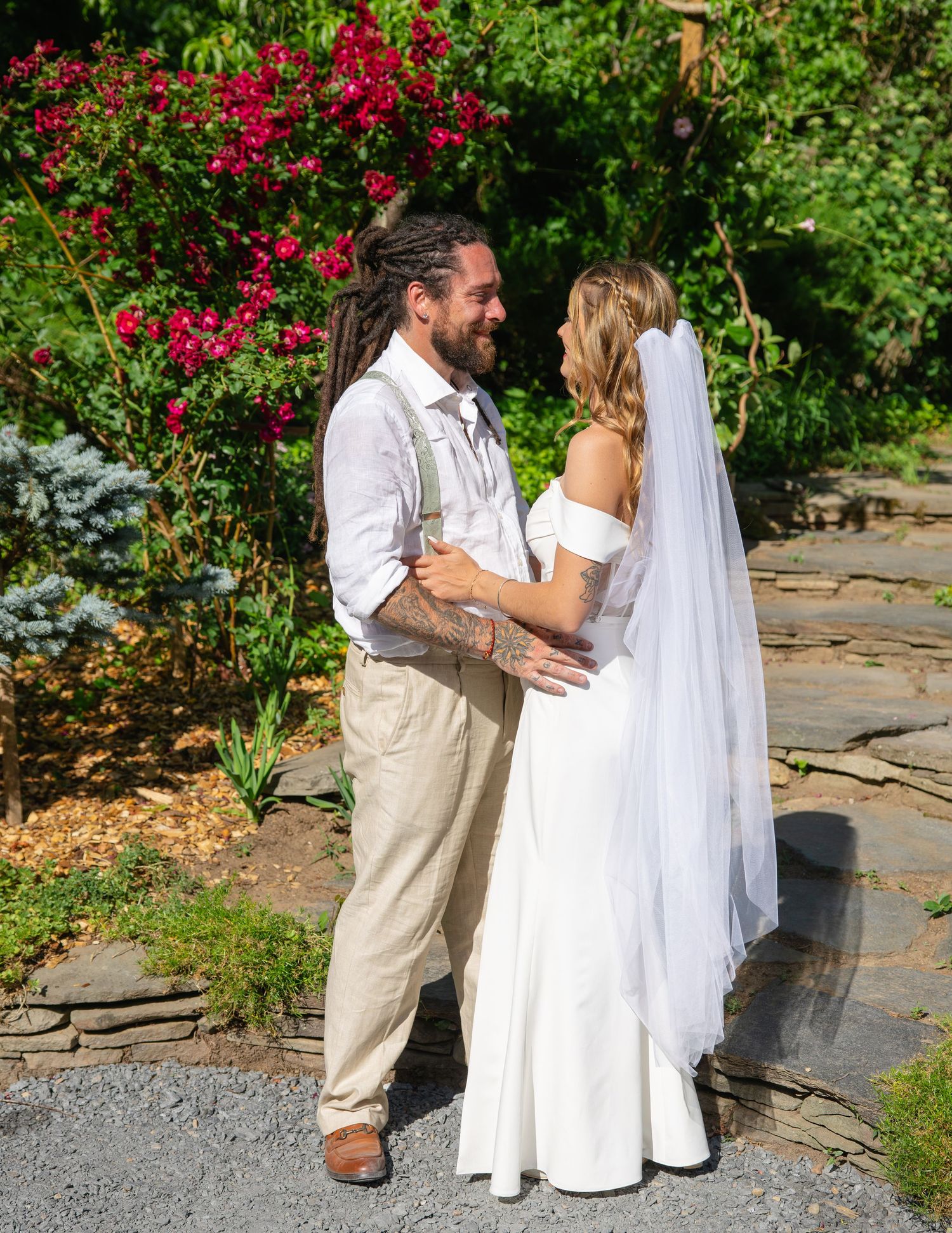 Newlyweds share a romantic moment in a garden surrounded by vibrant red flowers and green foliage on their wedding day.