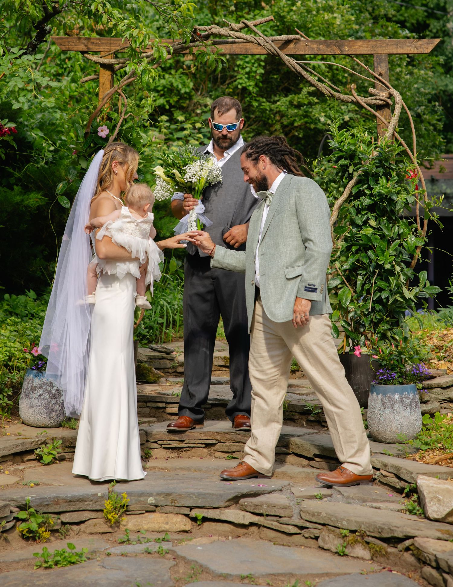 Outdoor garden wedding ceremony with bride holding baby, groom, and officiant under wooden arbor.