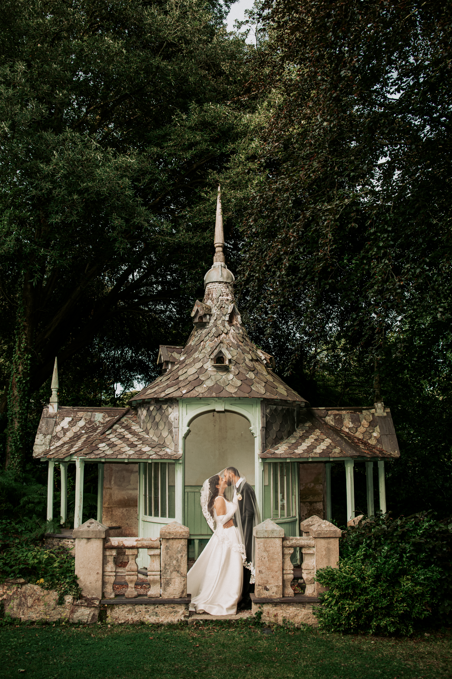 Bride and groom kissing in front of a charming vintage gazebo surrounded by lush green trees.