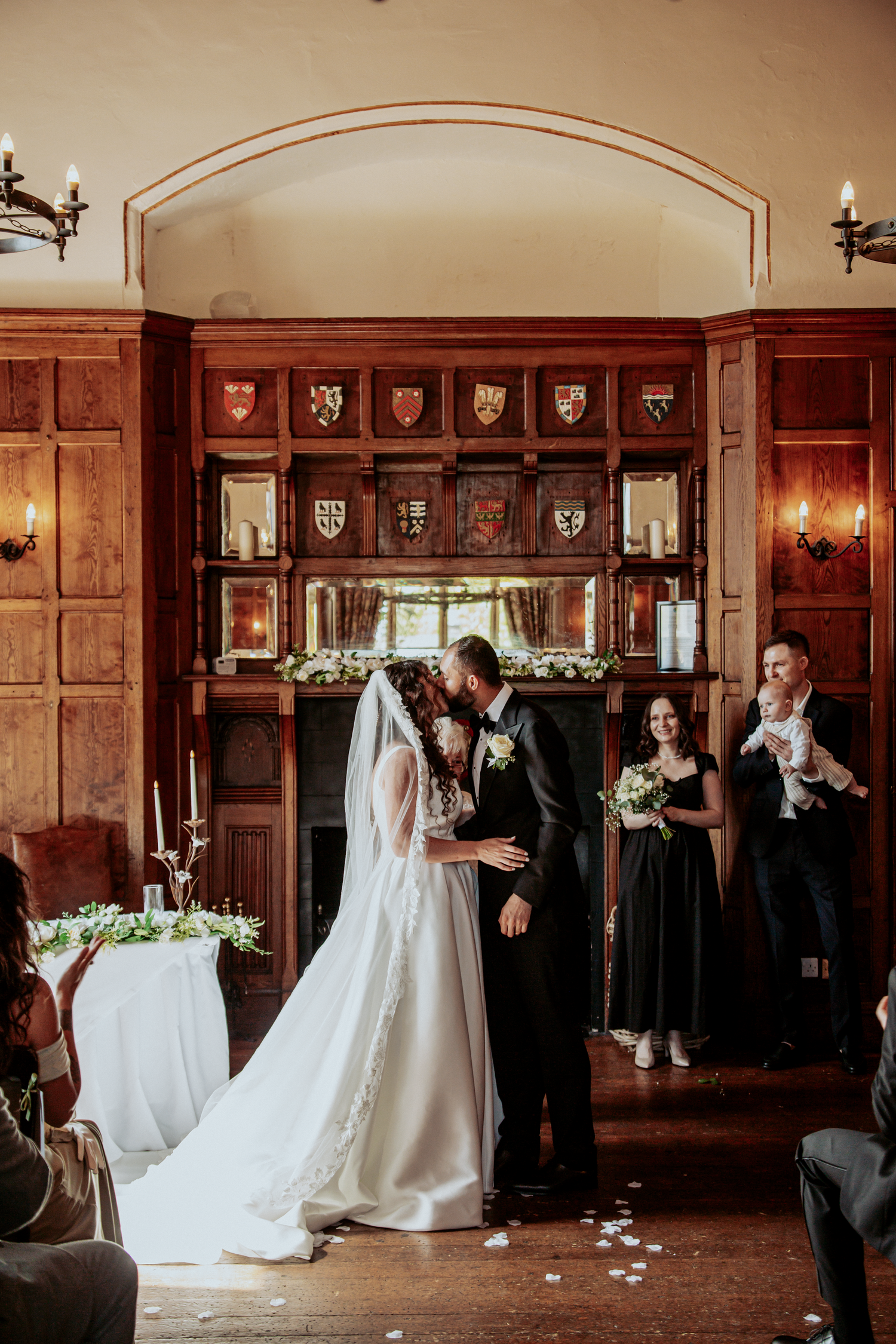Bride and groom share first kiss at elegant wood-paneled wedding ceremony with floral fireplace backdrop.