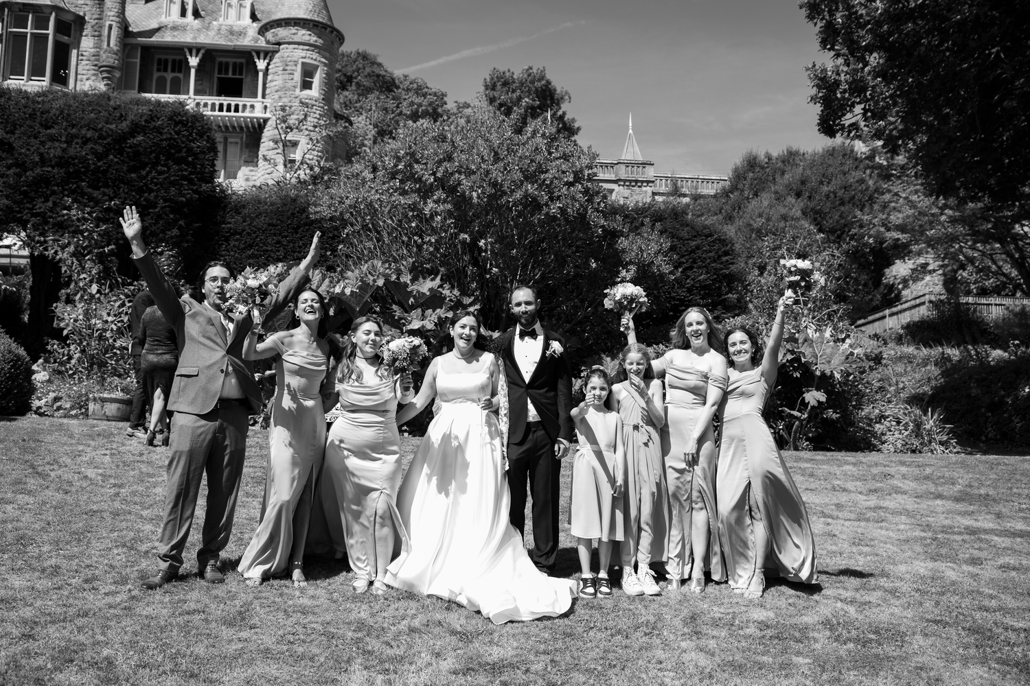 Black and white wedding party photo on lawn with bride, groom, bridesmaids, and guests celebrating outdoors.