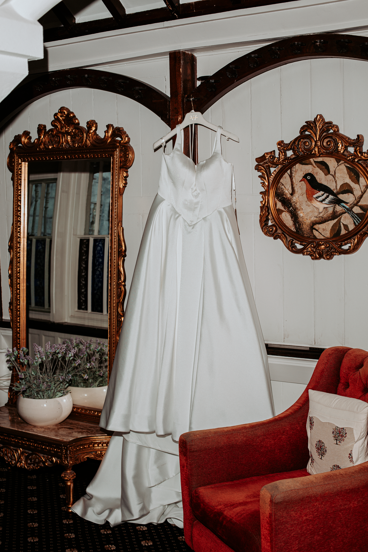 Elegant white satin wedding dress hanging in a vintage room with gold-framed mirrors and red velvet armchair.