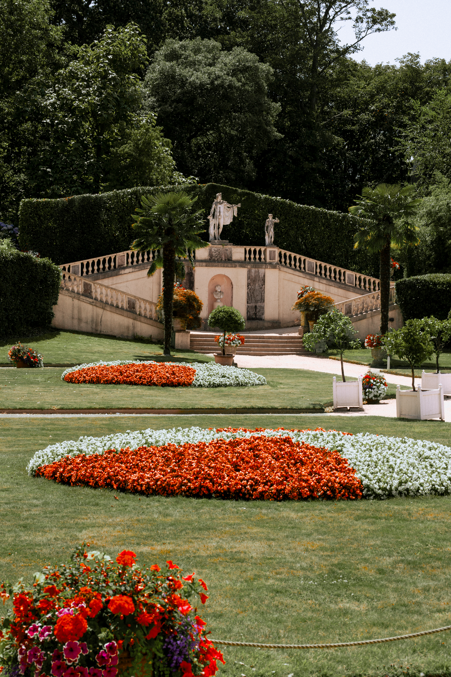 Formal Italian garden with colorful flower beds, manicured hedges, and classical statues on ornate staircase.