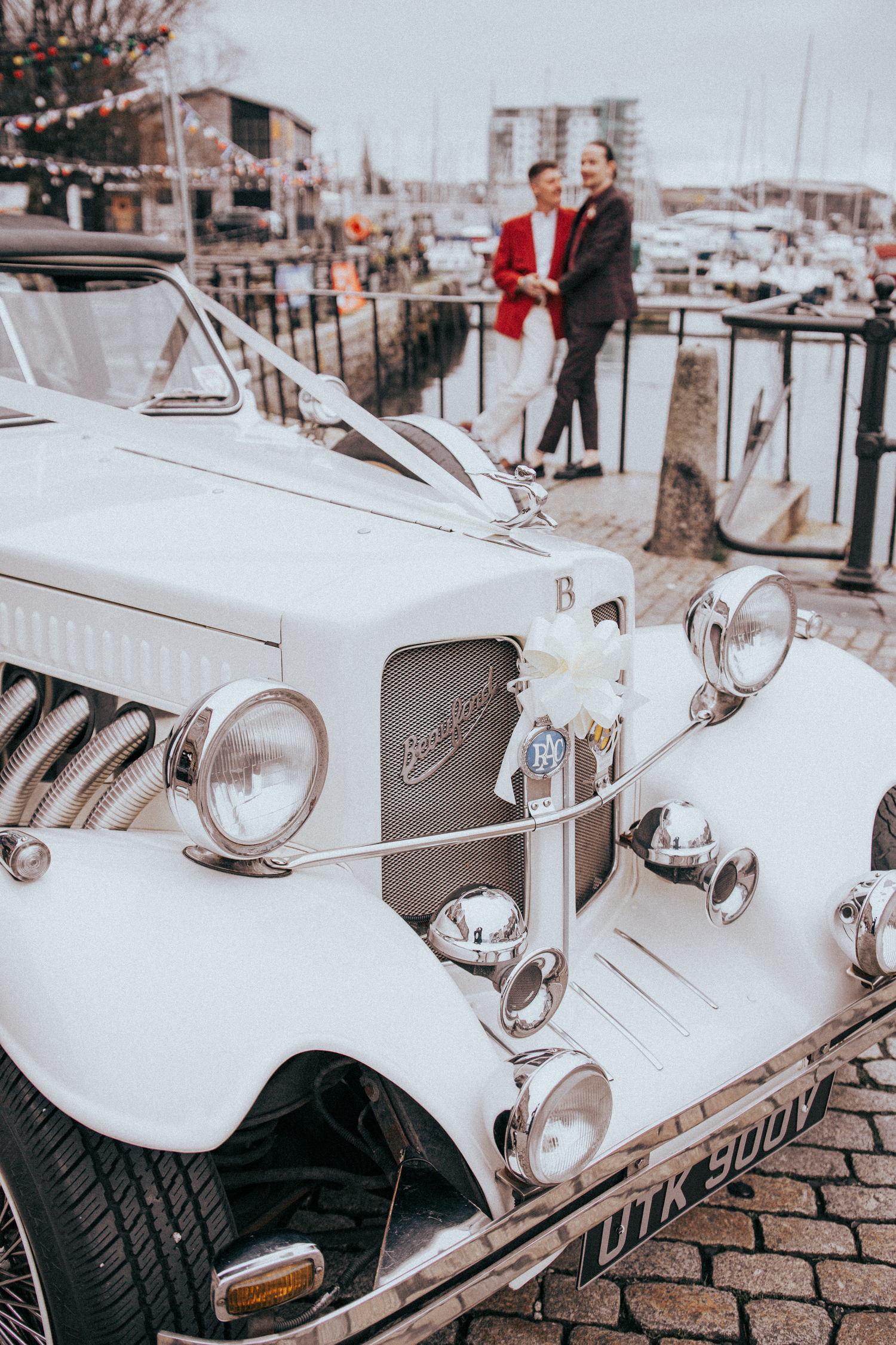 Two grooms in red and white suits stand by a classic white Beauford wedding car at a marina.