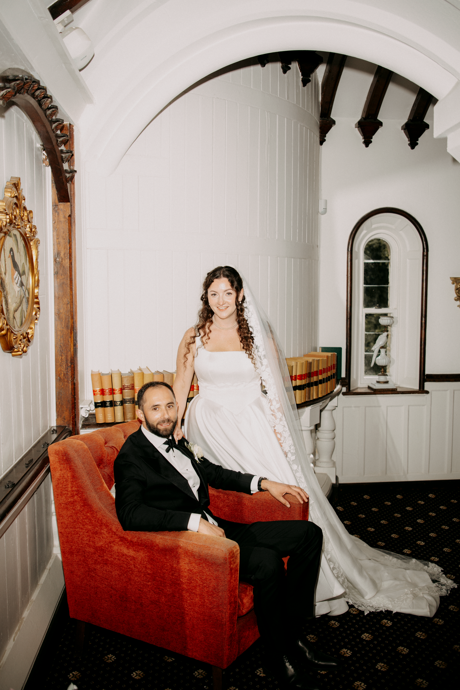 Bride in white gown stands behind groom in tuxedo seated in red velvet chair inside elegant venue.