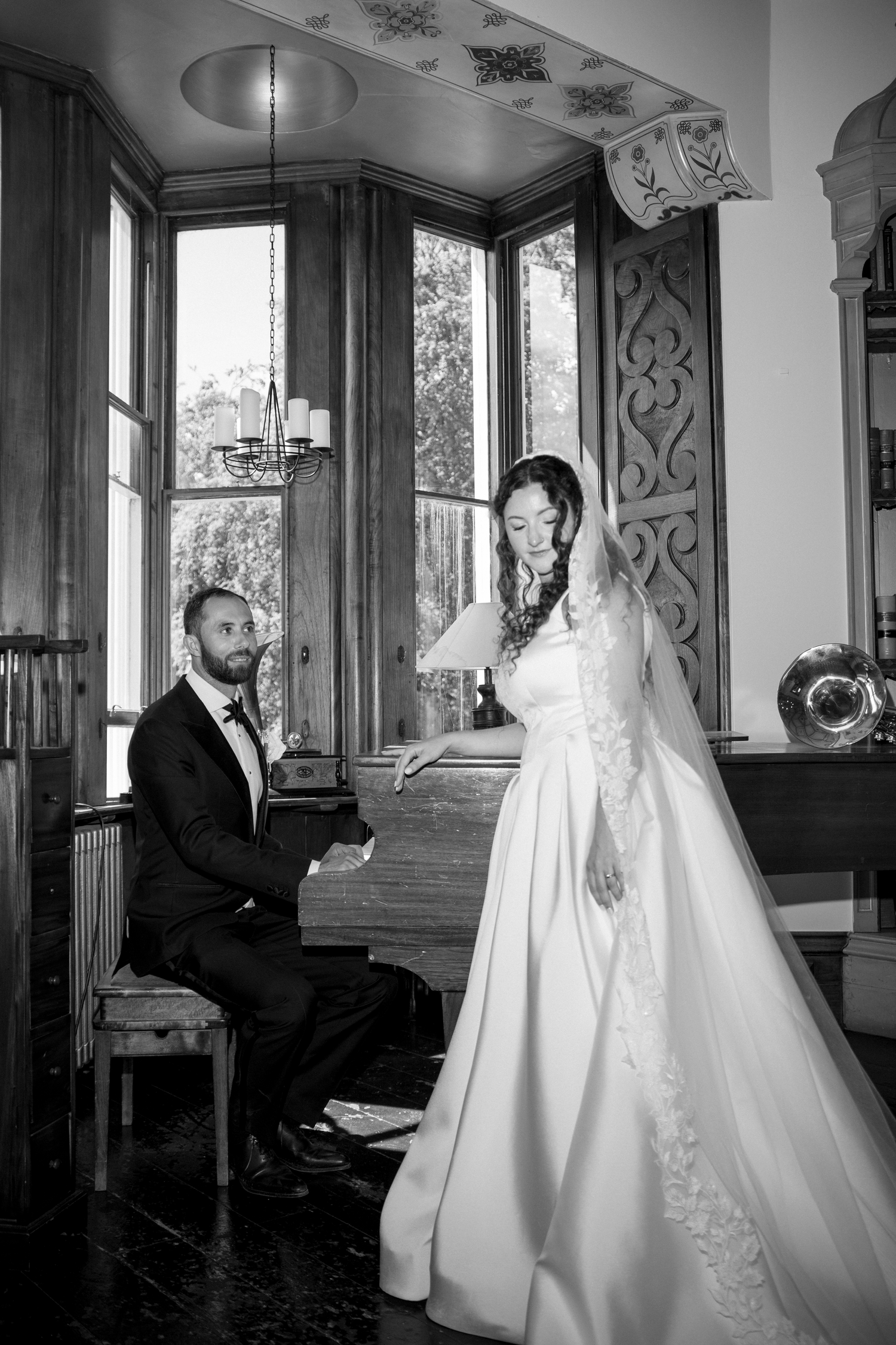 Black and white wedding photo of bride in gown standing by groom seated at piano in elegant room.
