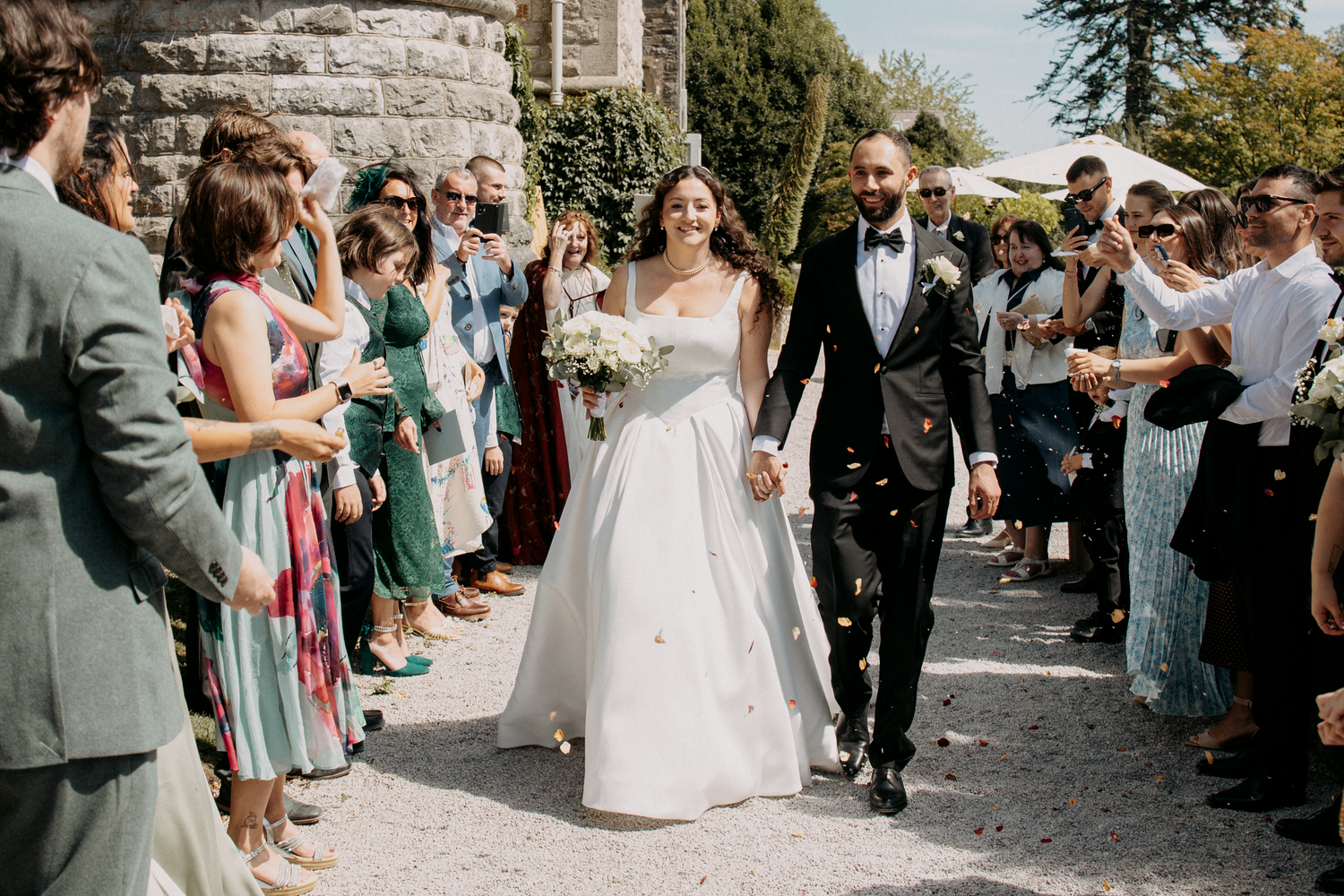 Bride and groom walk through cheering guests throwing confetti at outdoor wedding ceremony.