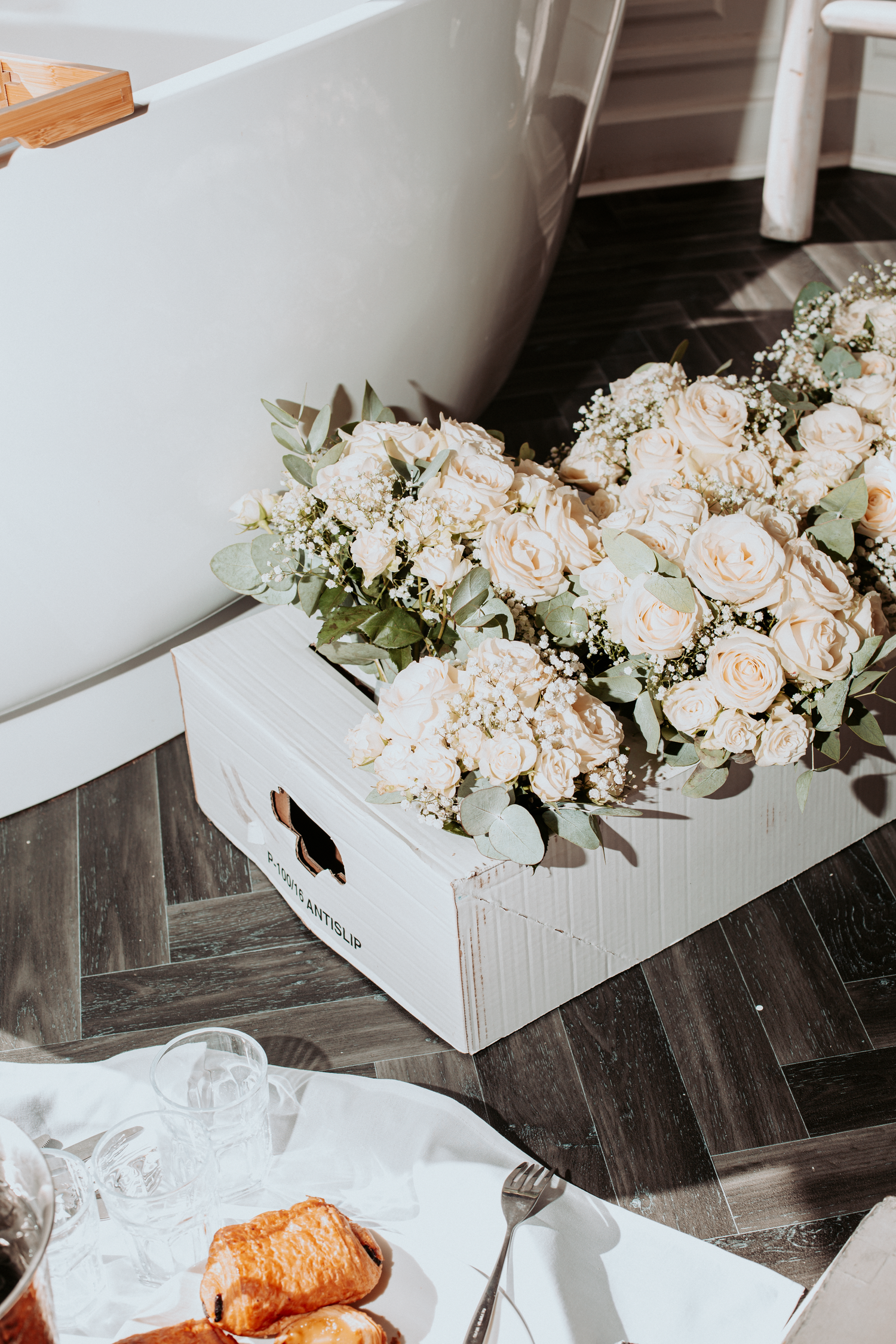 White roses and eucalyptus arrangement in a white box beside a bathtub with pastries and glasses nearby.