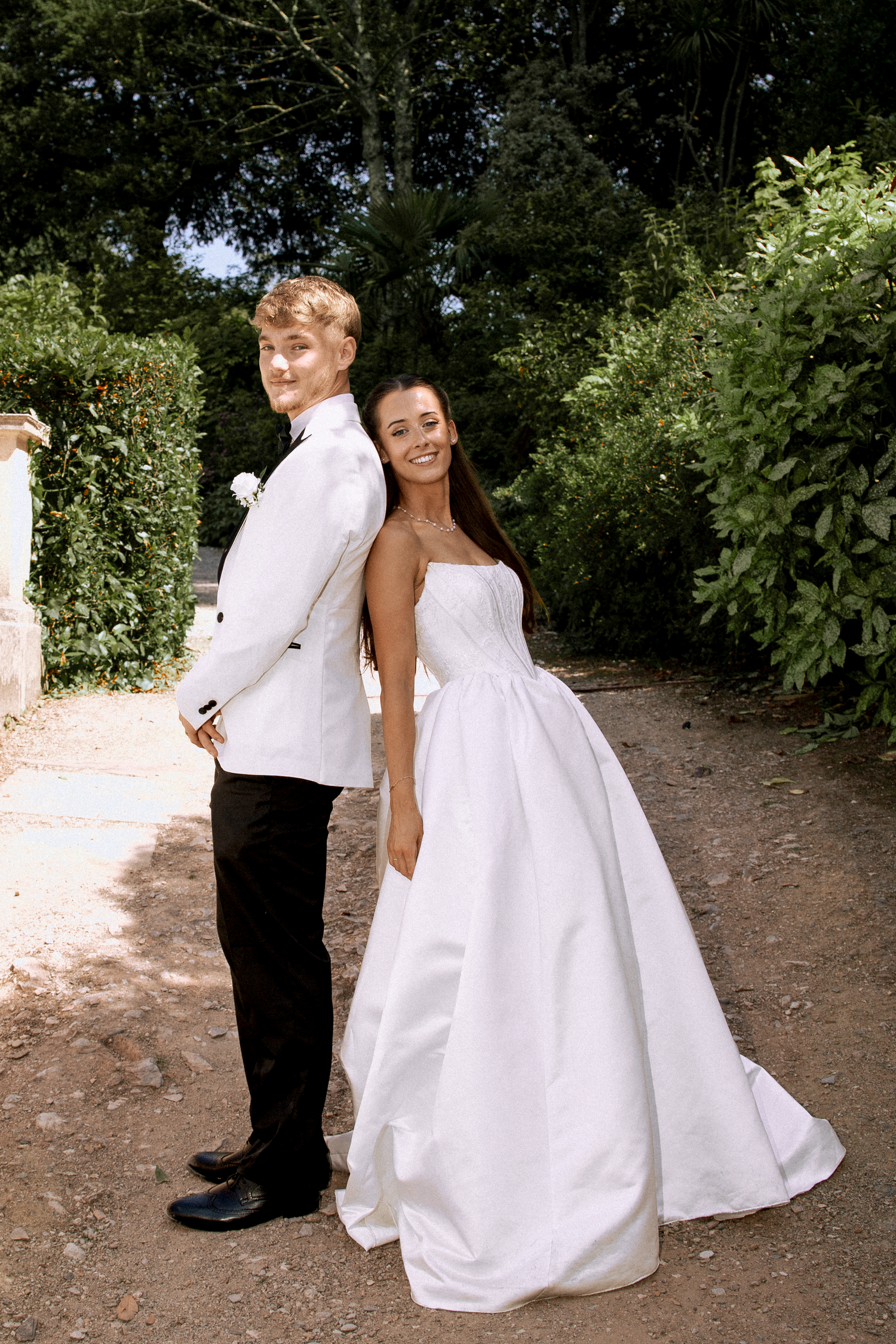 Bride in a strapless white ball gown and groom in white jacket standing back to back on a garden path.