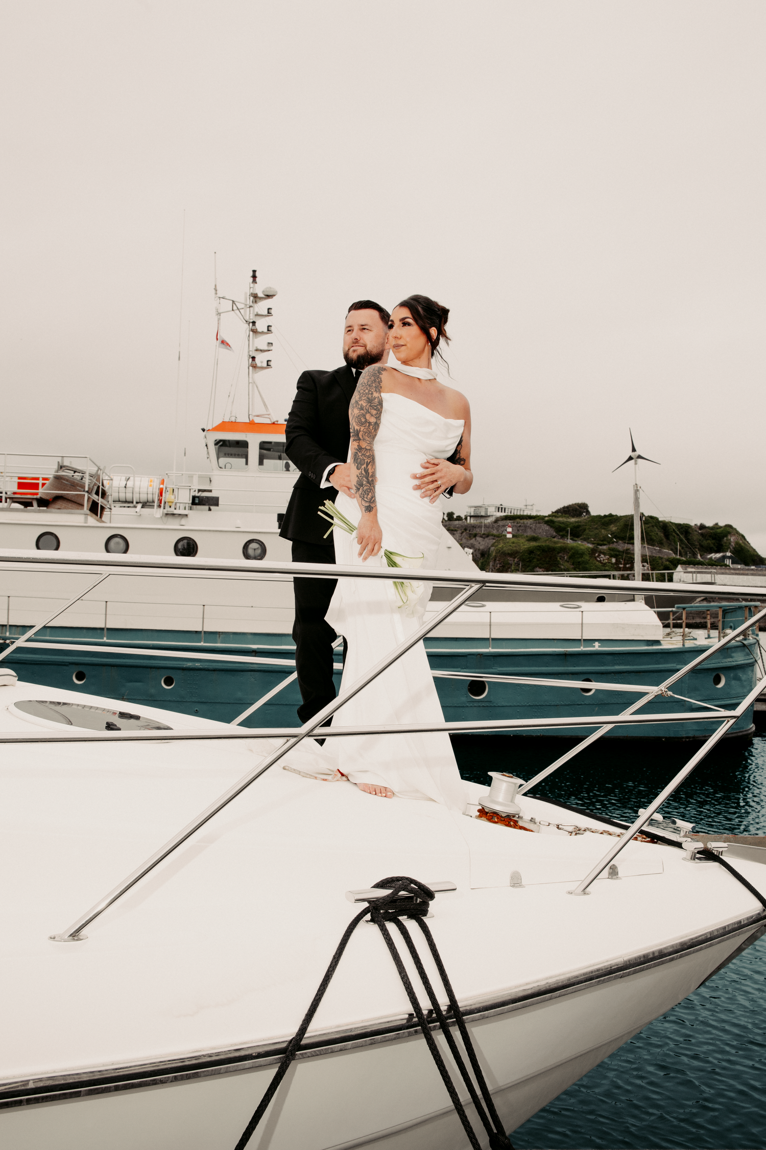 Wedding couple standing together on a white yacht deck at a harbor, bride in sleek gown with tattoos.