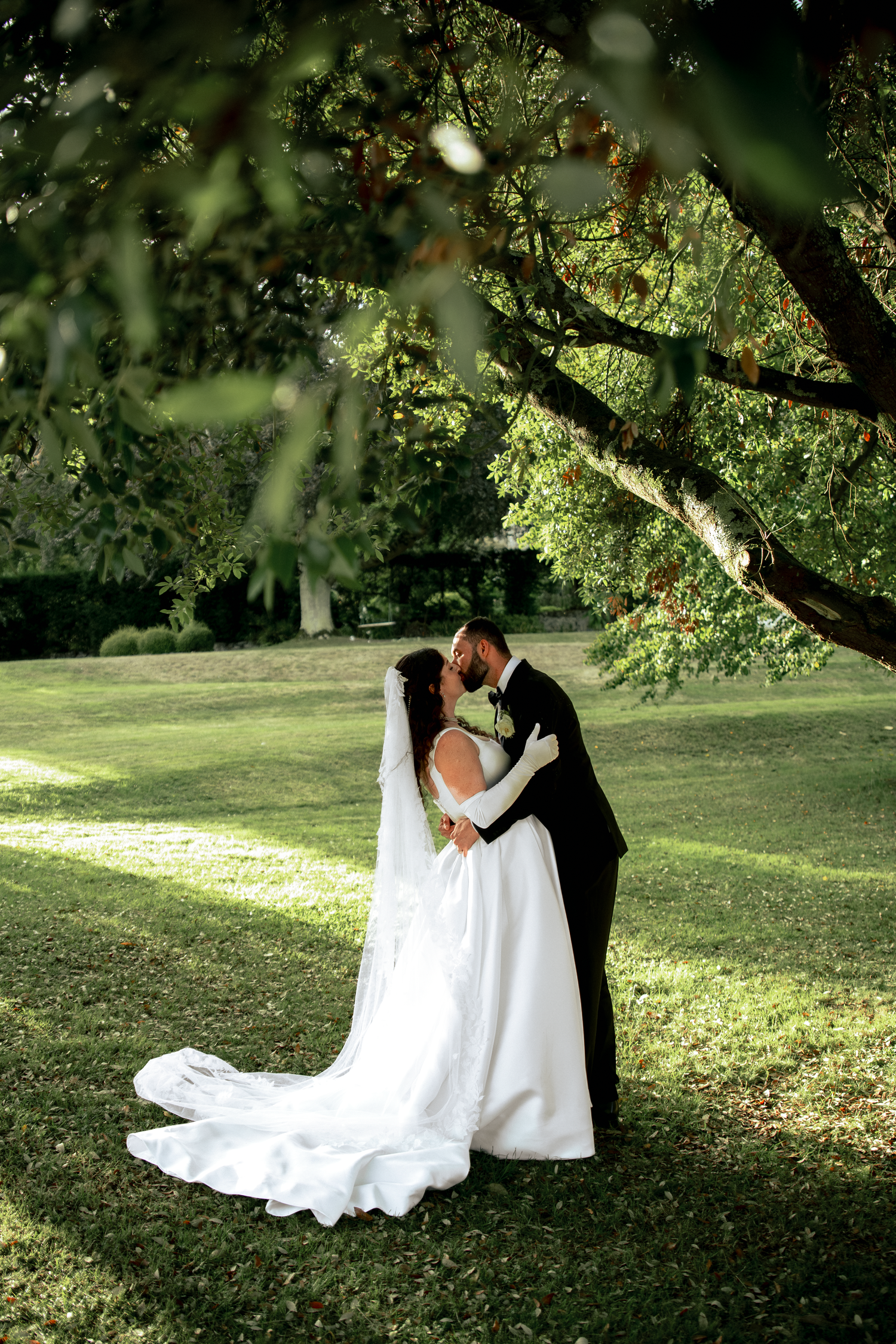 Bride and groom share a romantic kiss under lush green trees on a sunlit lawn, bride wearing a flowing white gown.