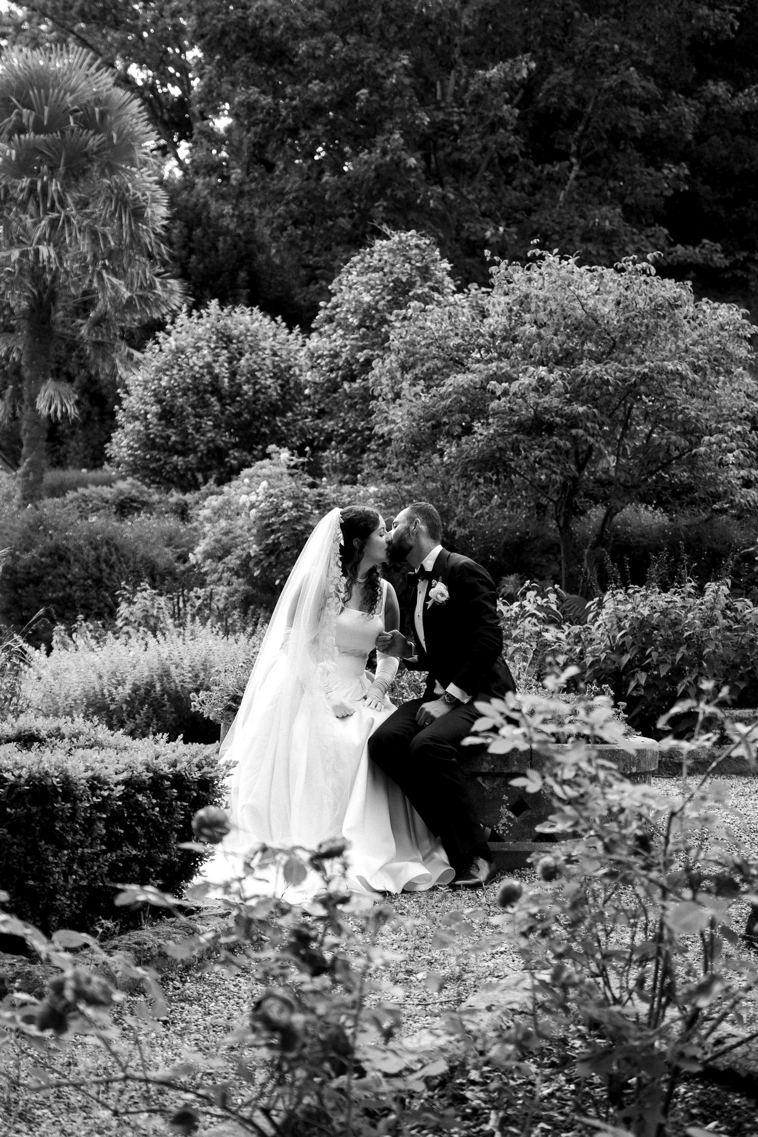 Black and white photo of a bride and groom sharing a kiss while seated in a lush garden on their wedding day.