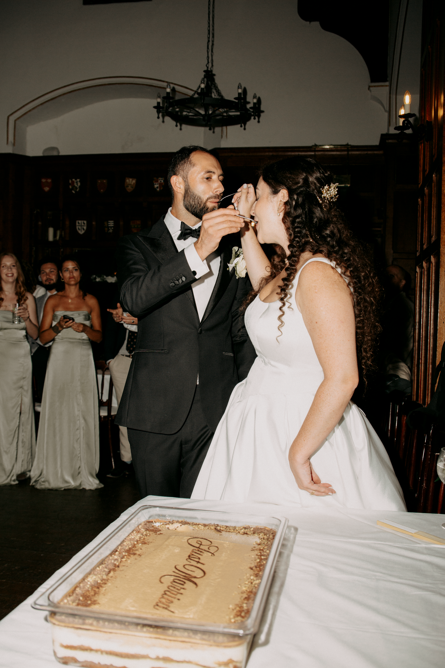 Bride and groom share a cake tasting moment at their elegant wedding reception with guests seated in background.