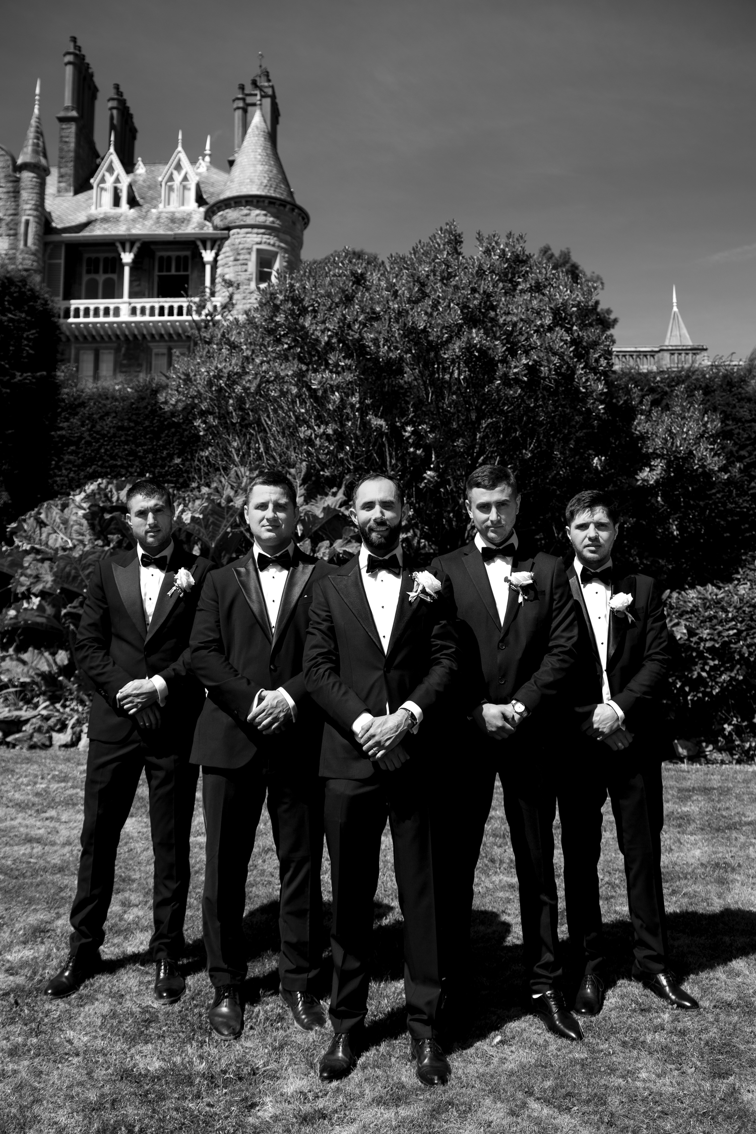 Five groomsmen in black tuxedos with bow ties pose outdoors before a grand castle-style building in a black and white photo.
