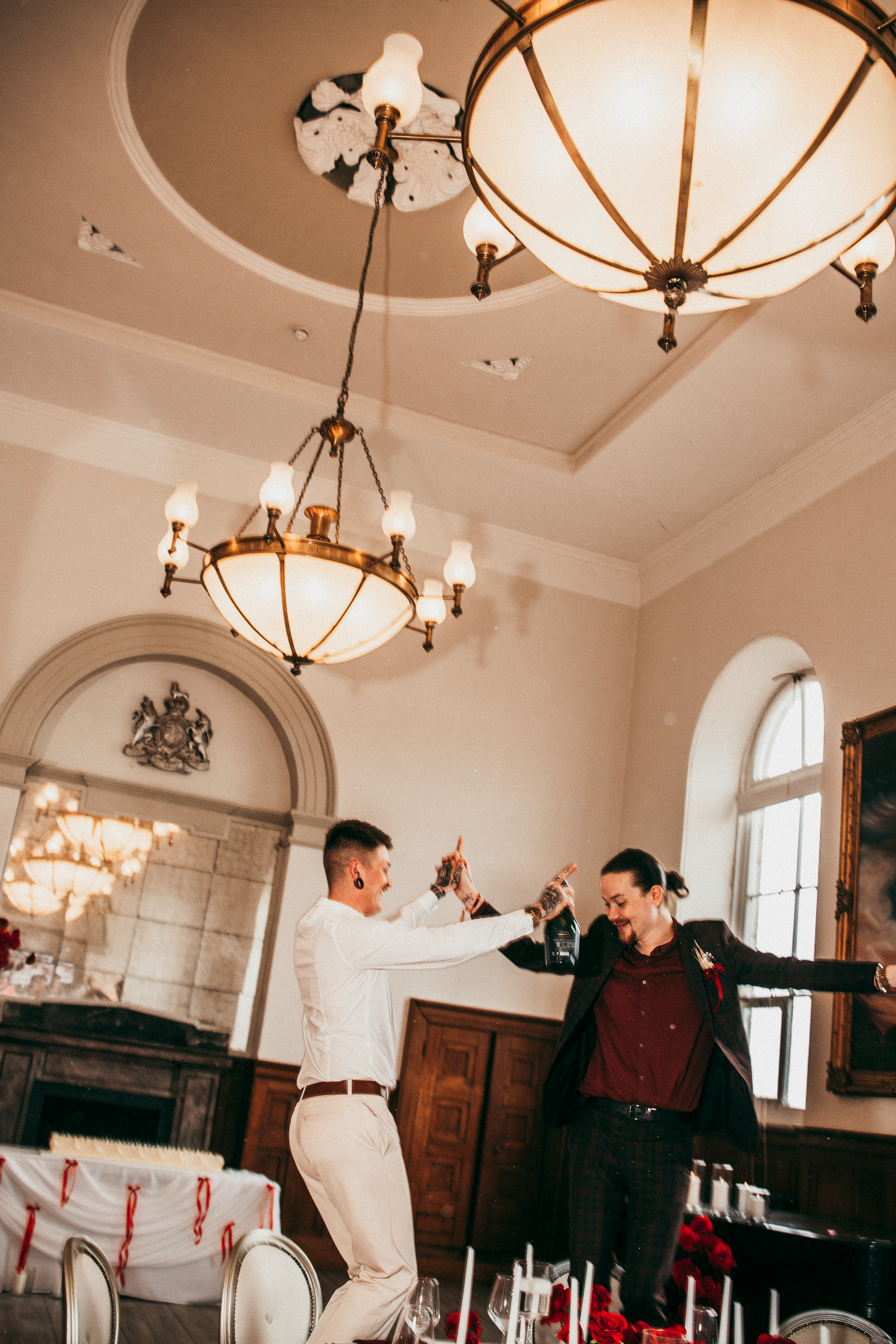 Two grooms dancing together under ornate chandelier in elegant wedding reception ballroom.