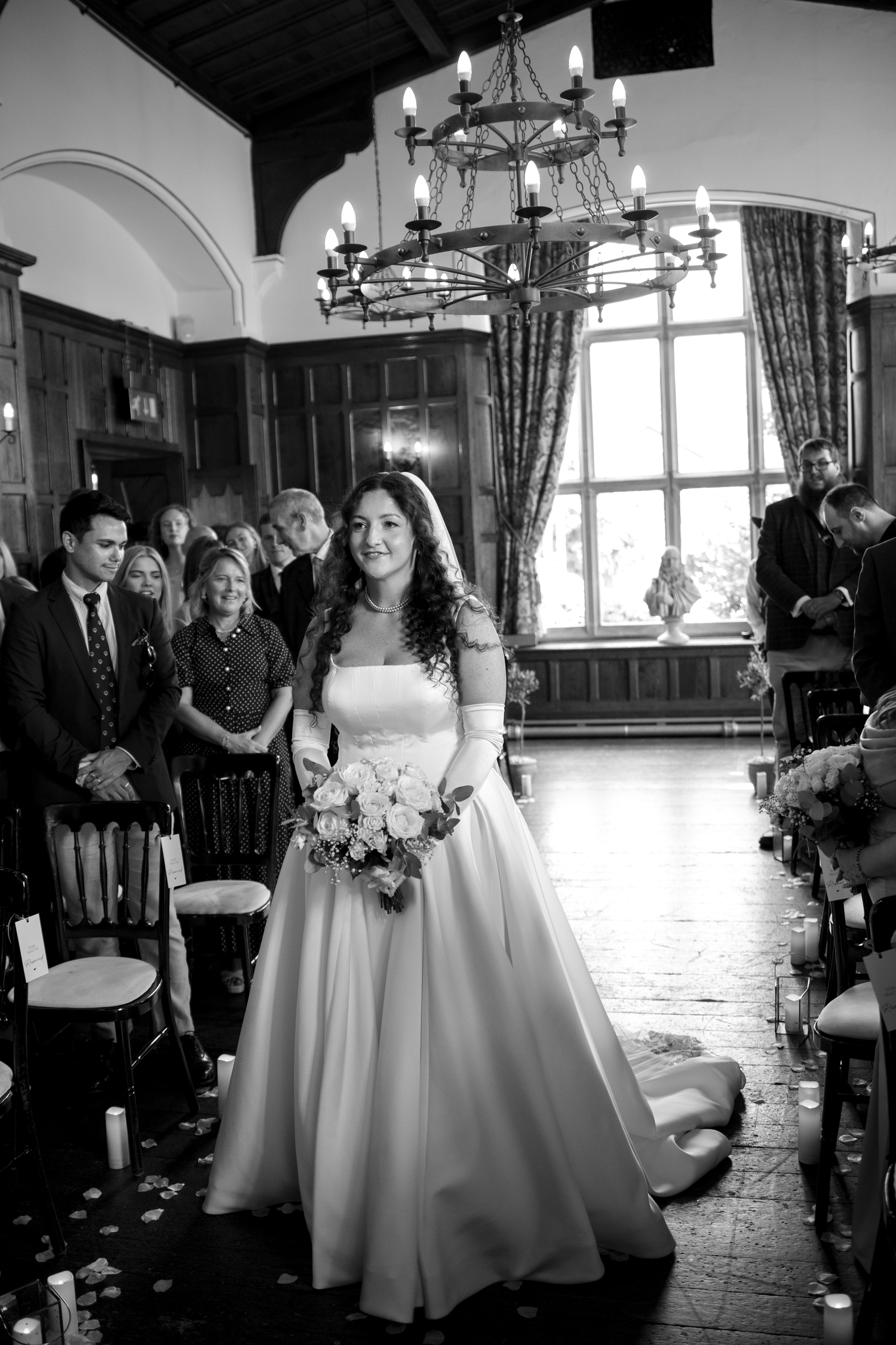 Black and white photo of a bride walking down the aisle in a ballgown, holding a bouquet, with guests seated in a grand hall.