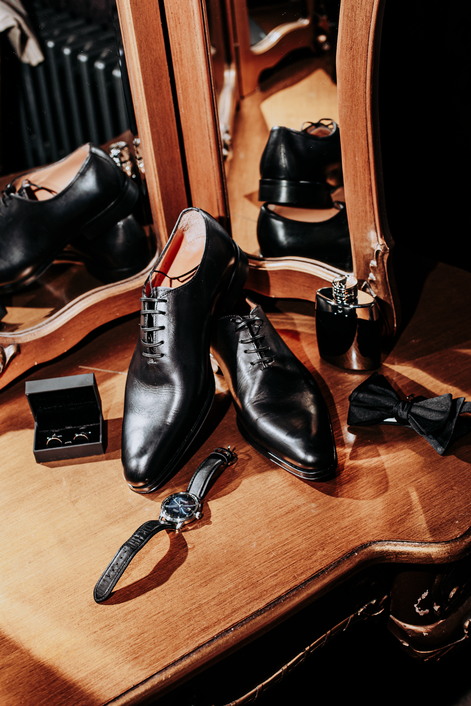Black leather oxford shoes with a watch, bow tie, and cufflinks on a wooden dresser with mirror reflection.