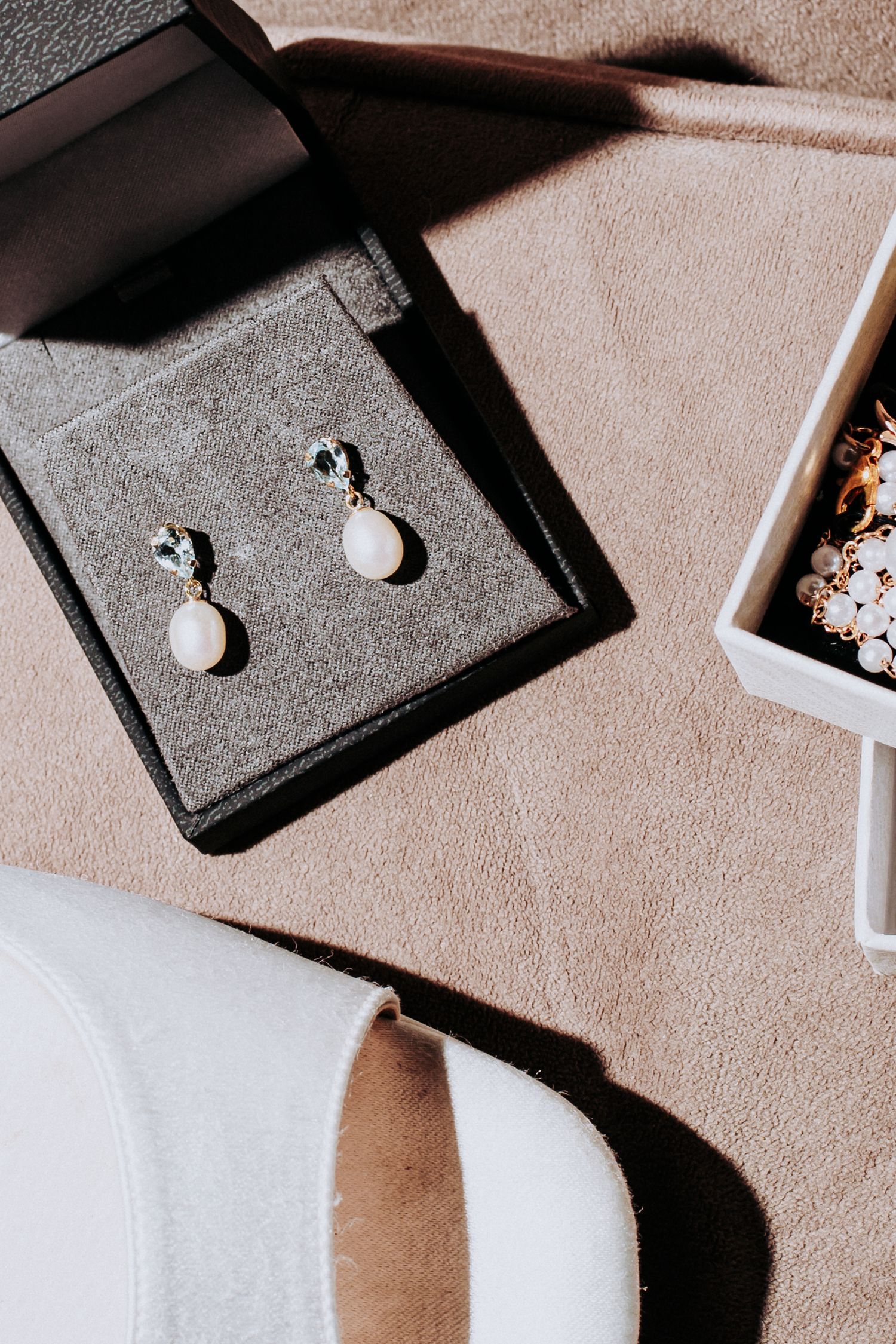 Elegant pearl drop earrings in a gray velvet jewelry box on a beige suede surface with accessories nearby.