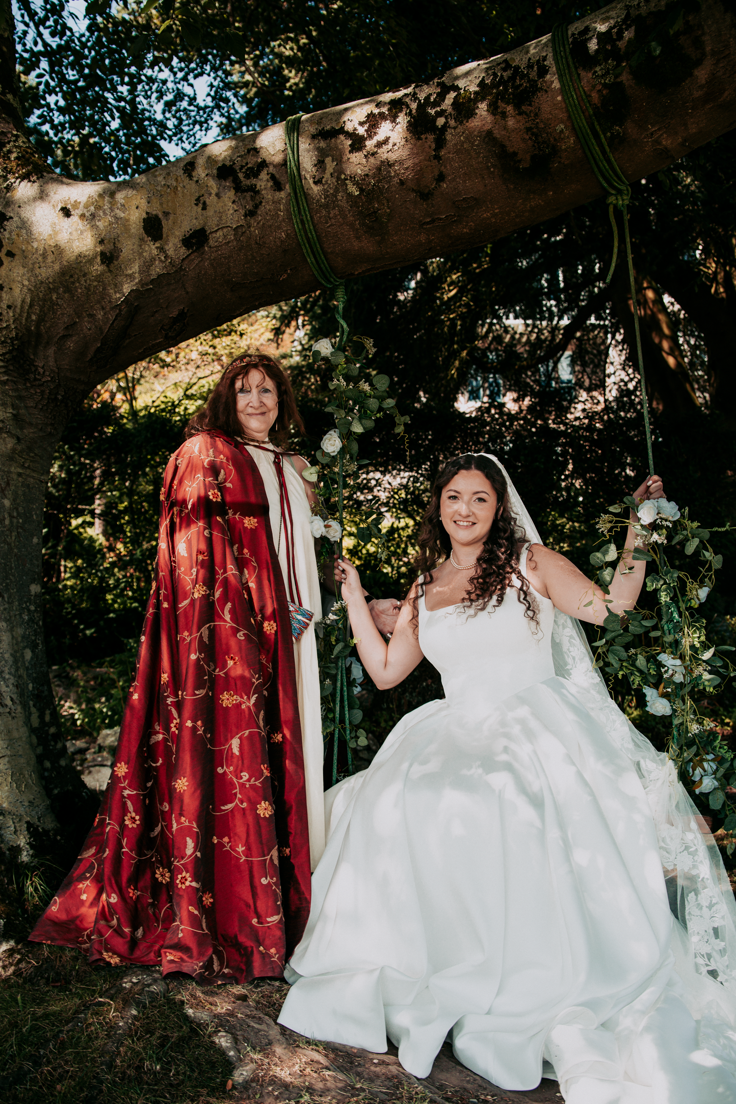 Bride in white gown sits on swing beside woman in red embroidered cloak under a tree.