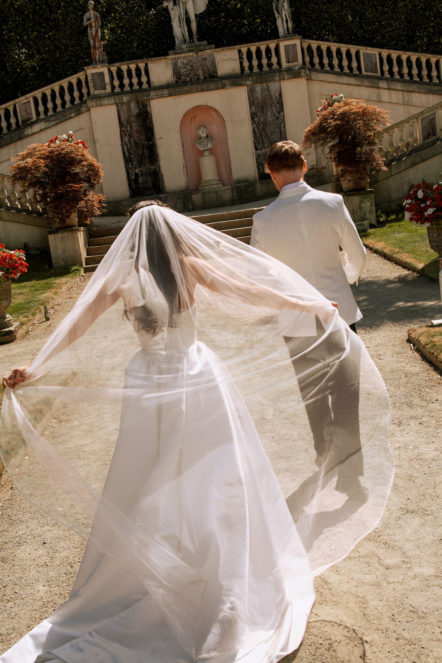 Bride's veil flowing dramatically as couple walks toward ornate garden staircase on wedding day.