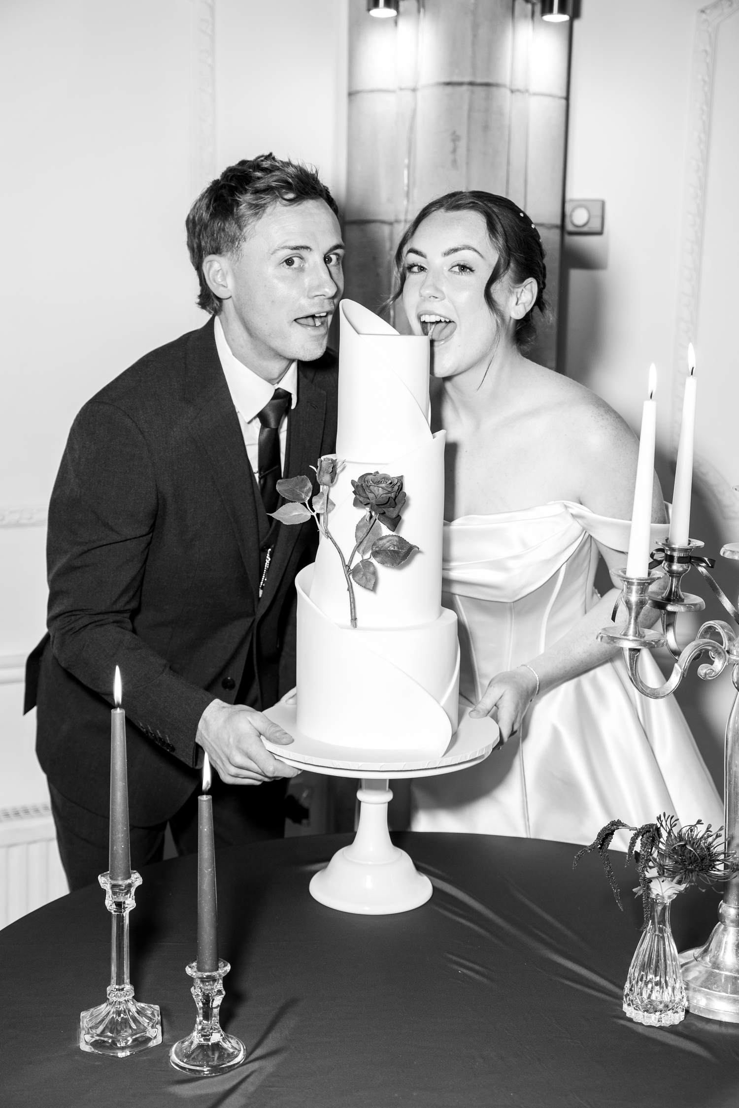 Black and white photo of a bride and groom playfully pretending to eat a tall white wedding cake with rose decorations.