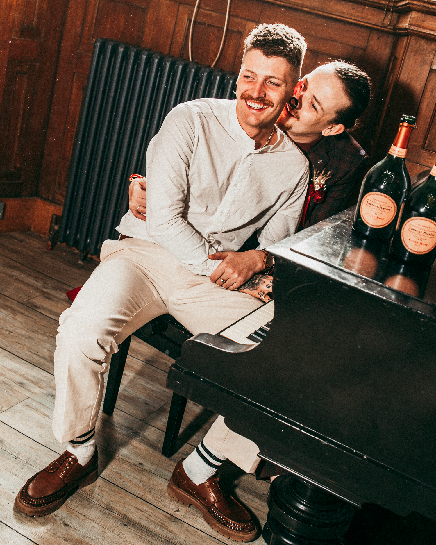Two grooms sitting at black piano laughing together with champagne bottles at wedding reception.