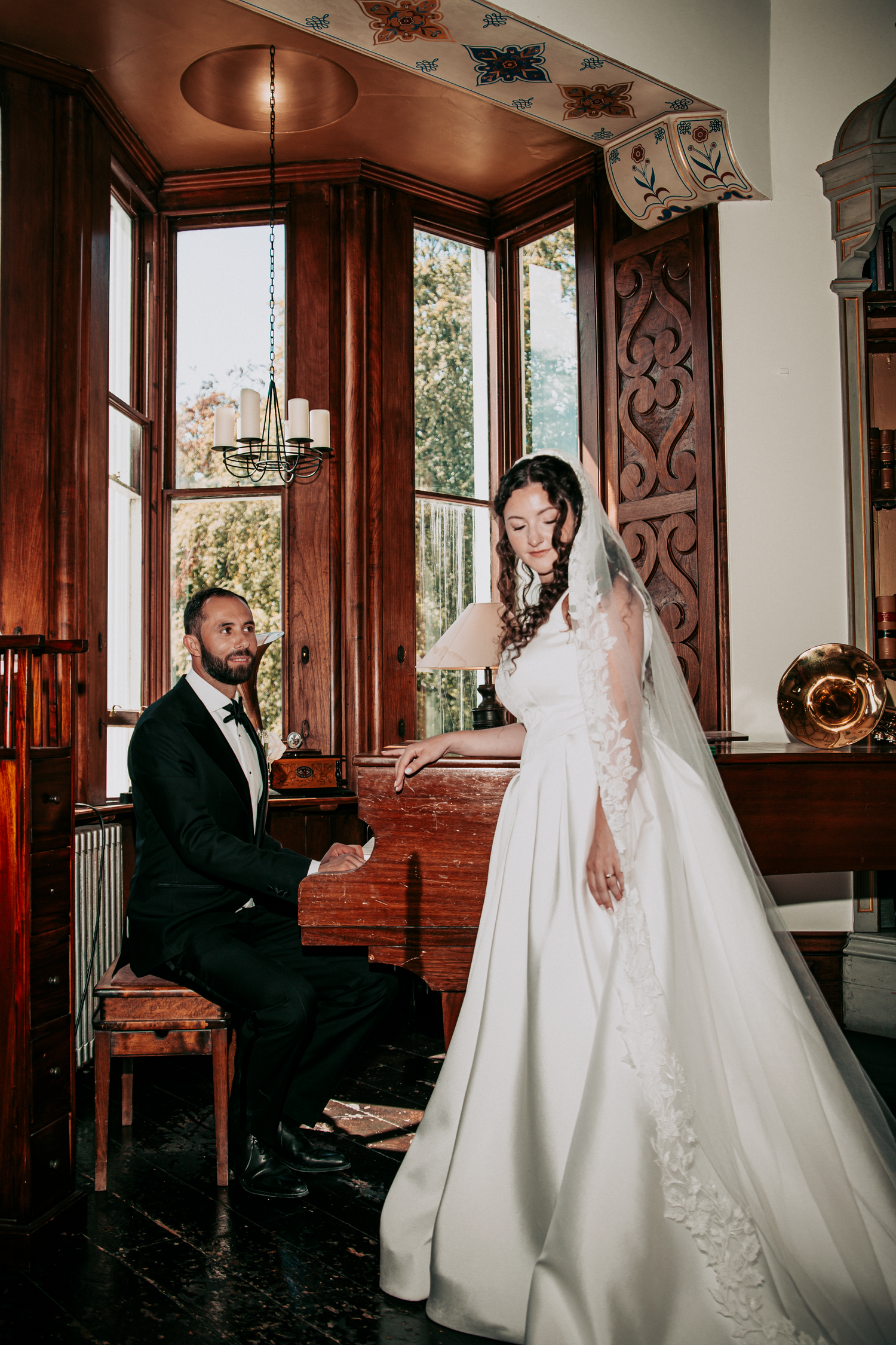 A bride in a white gown stands near a groom seated at a piano in an elegant wood-paneled room.