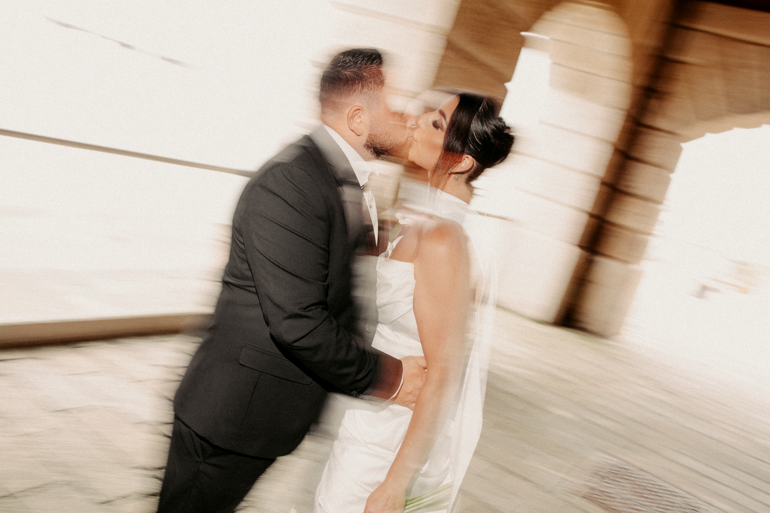 Bride and groom share a romantic blurred motion kiss in wedding attire near stone columns.