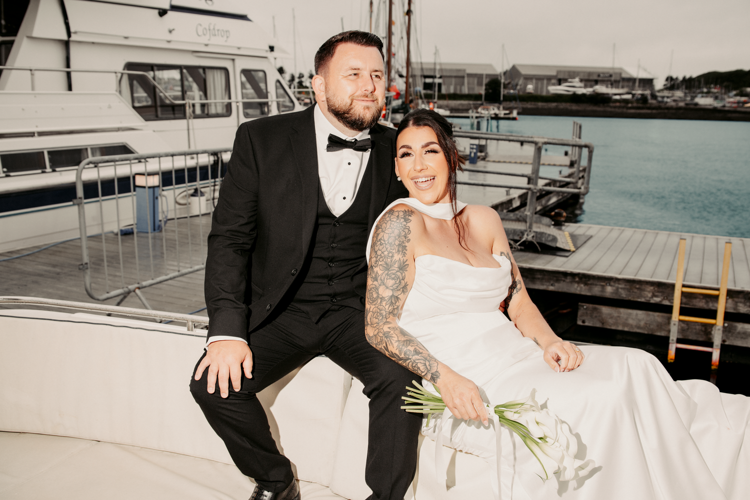 Smiling bride and groom sitting together on a yacht at the marina, bride holding green bouquet.