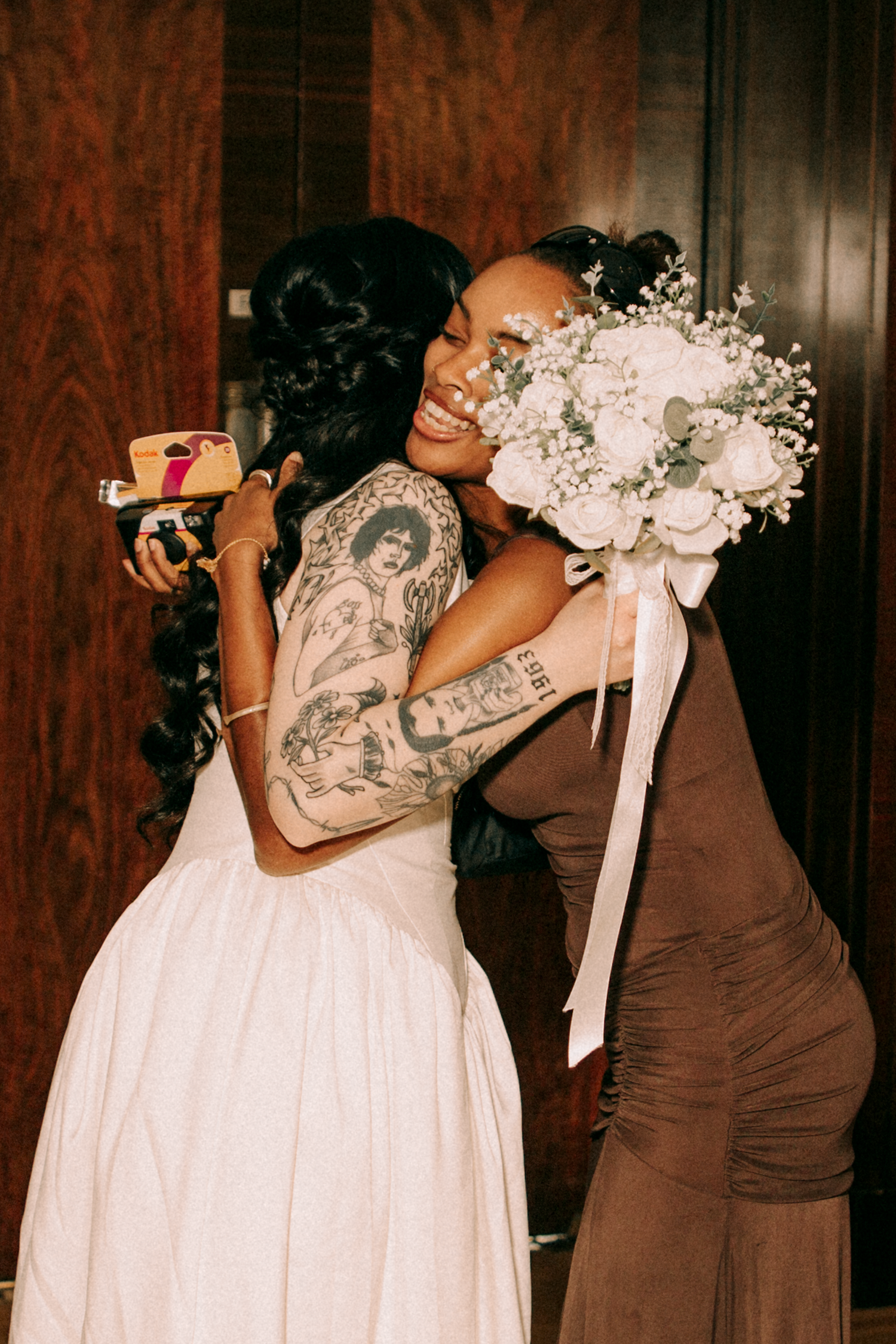 Tattooed bride in white dress hugging a bridesmaid in brown gown while holding a white floral bouquet.