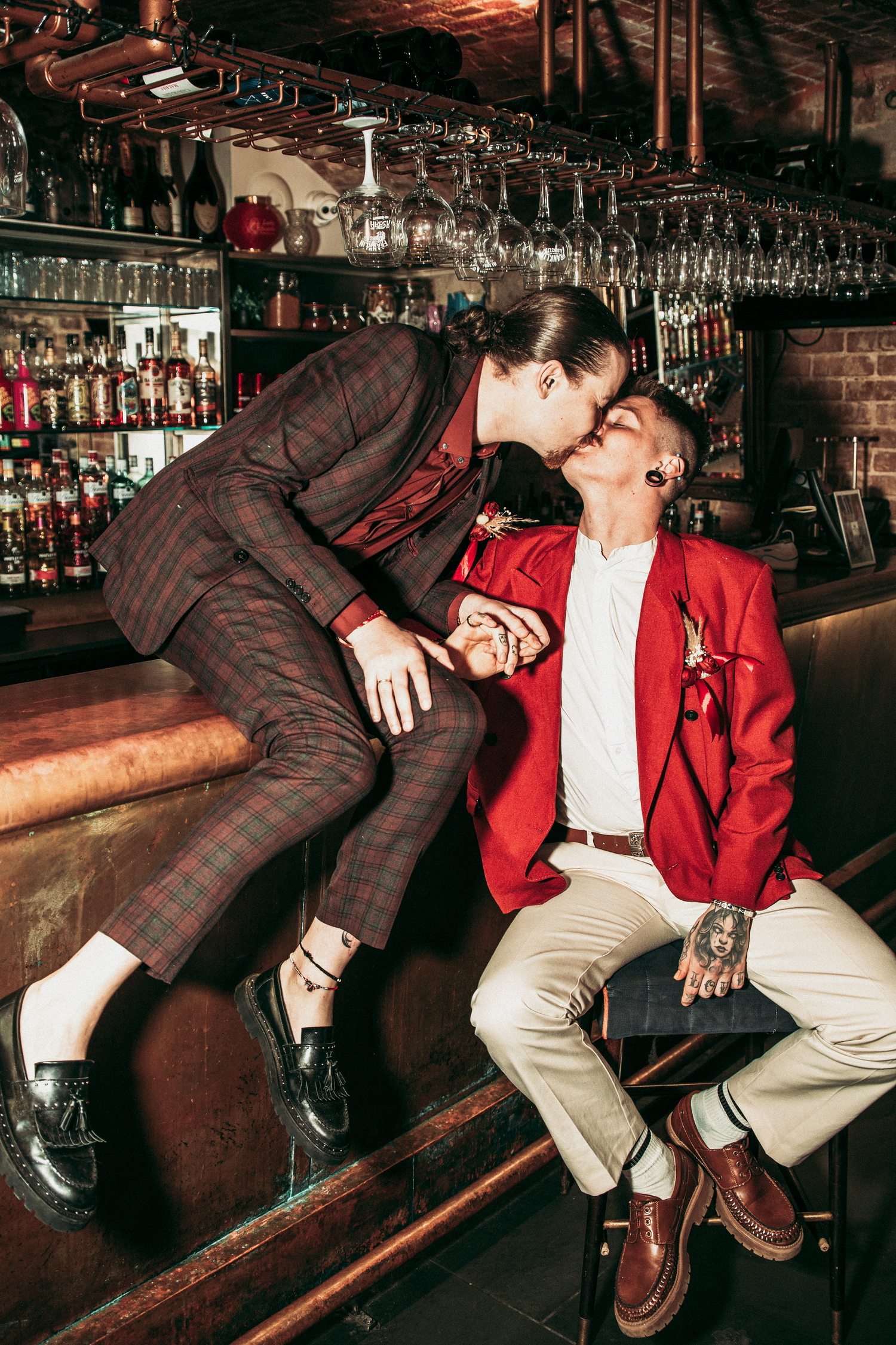 Two grooms share a kiss at a bar, one in plaid suit and one in a red blazer, holding hands.