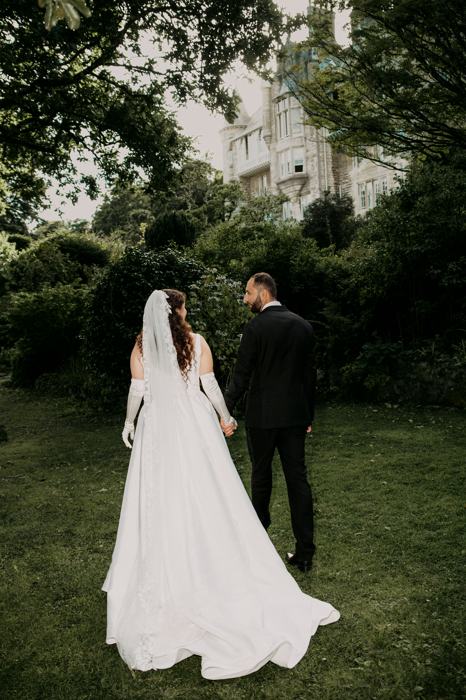 Bride and groom holding hands walking toward a grand estate surrounded by lush green trees on their wedding day.