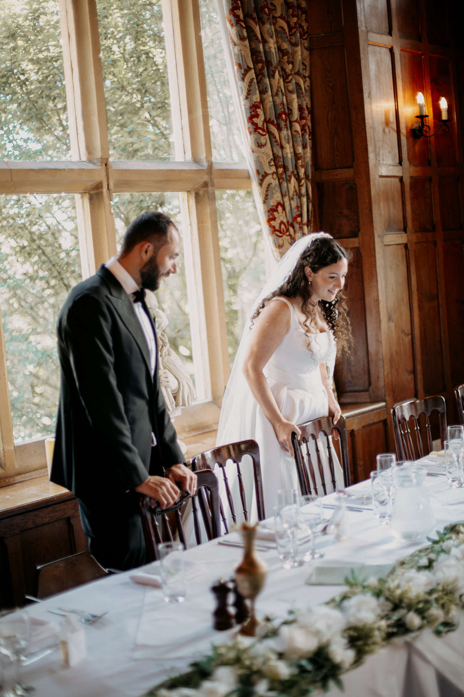 Bride and groom stand by elegantly set wedding reception table in a wood-paneled venue with large windows.